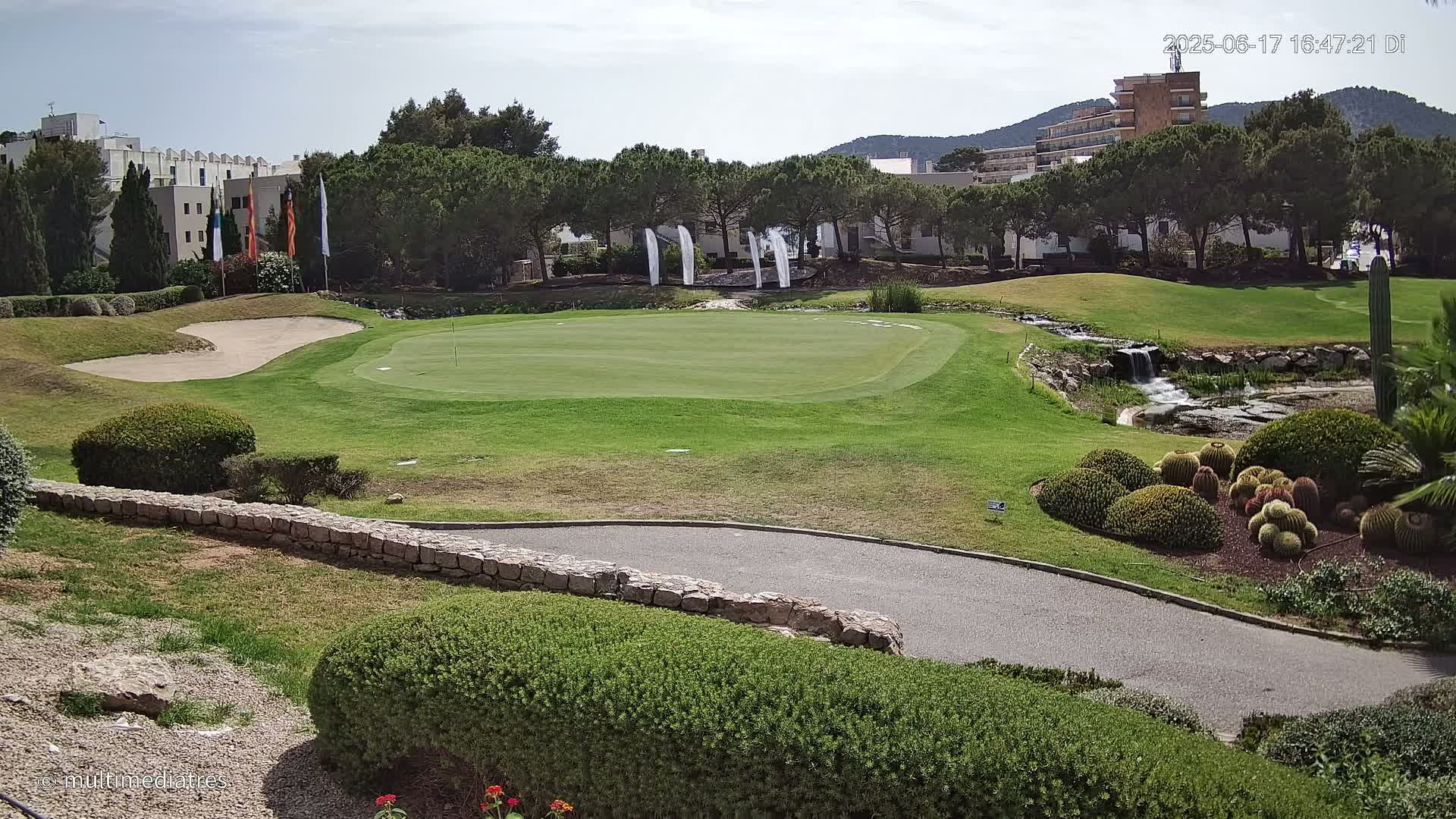 A golf course putting green is shown with a small stream and landscaping, under a mostly sunny sky.
