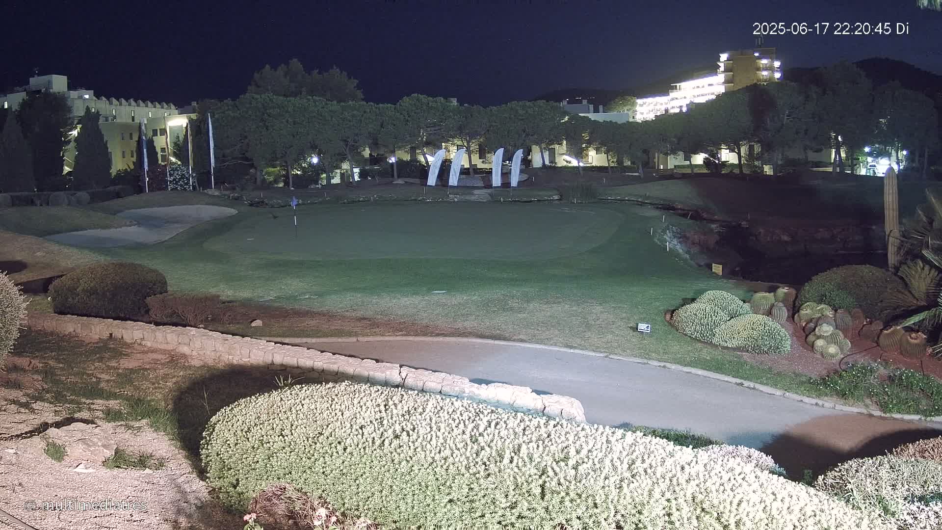 A nighttime view of a putting green at a golf course, illuminated by artificial light, with hotels and trees in the background under a clear, dark sky.