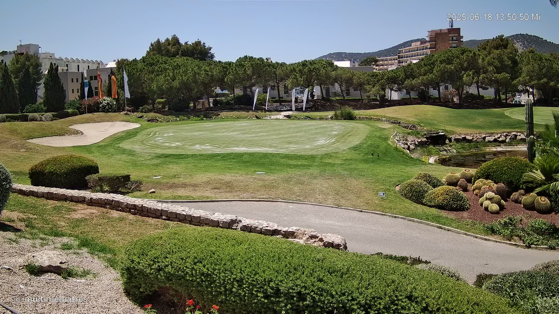 A putting green on a golf course with a sand trap, landscaping, and buildings in the background on a sunny day.