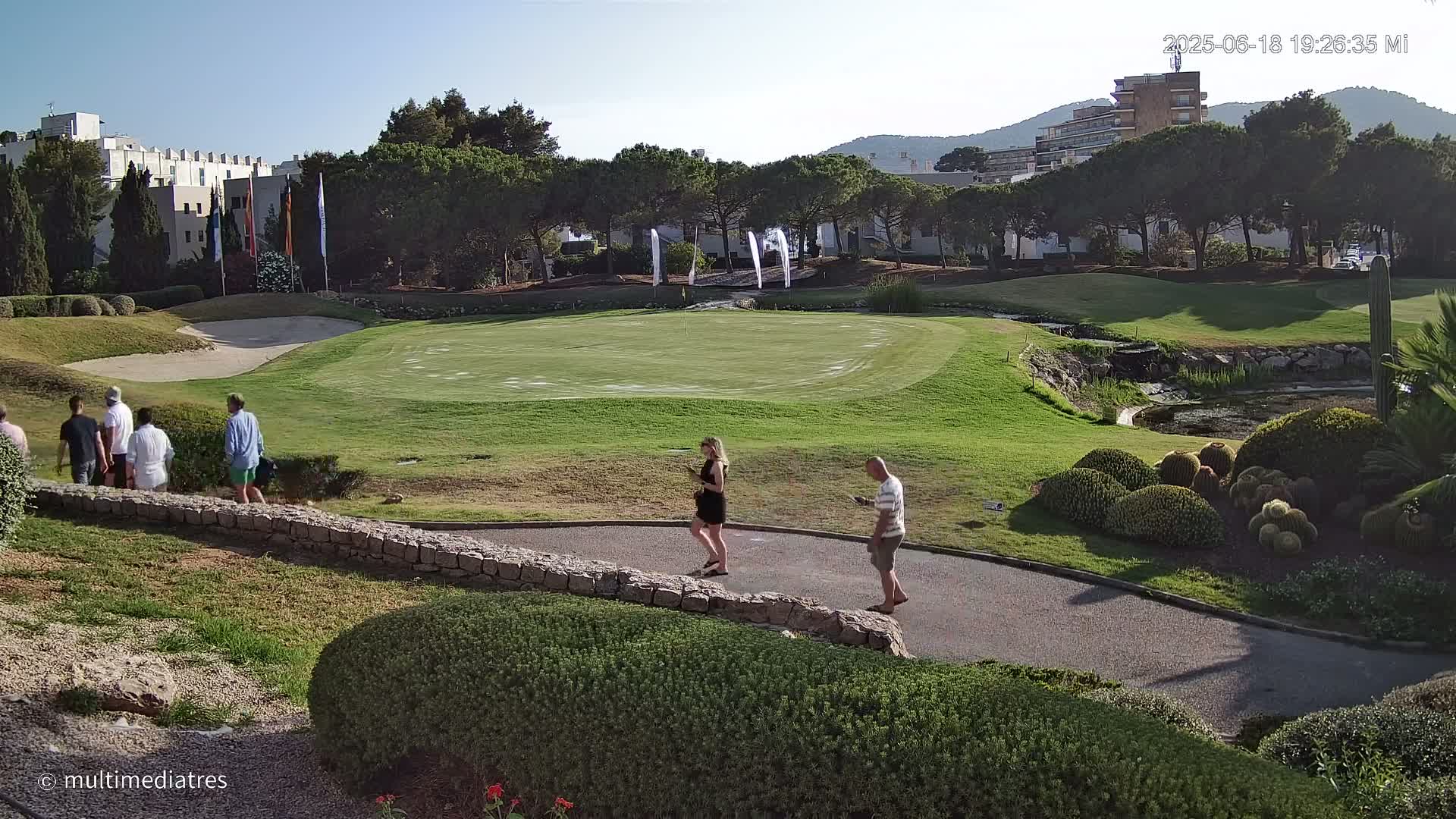 A golf course putting green with several people walking on a paved path, under a clear, sunny sky.