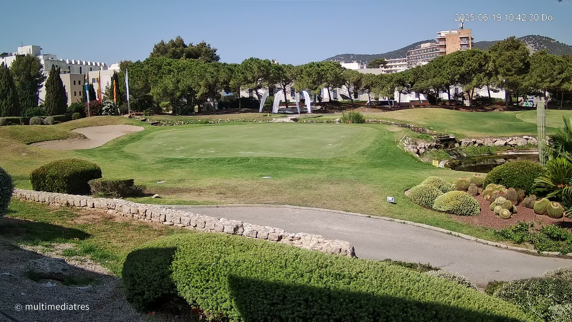 A putting green on a golf course is nestled among manicured lawns, landscaping, and buildings under a clear, sunny sky.