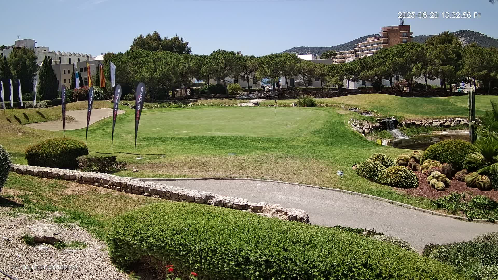A putting green on a golf course sits under a clear, sunny sky, surrounded by landscaping, a small waterfall, and buildings in the background.