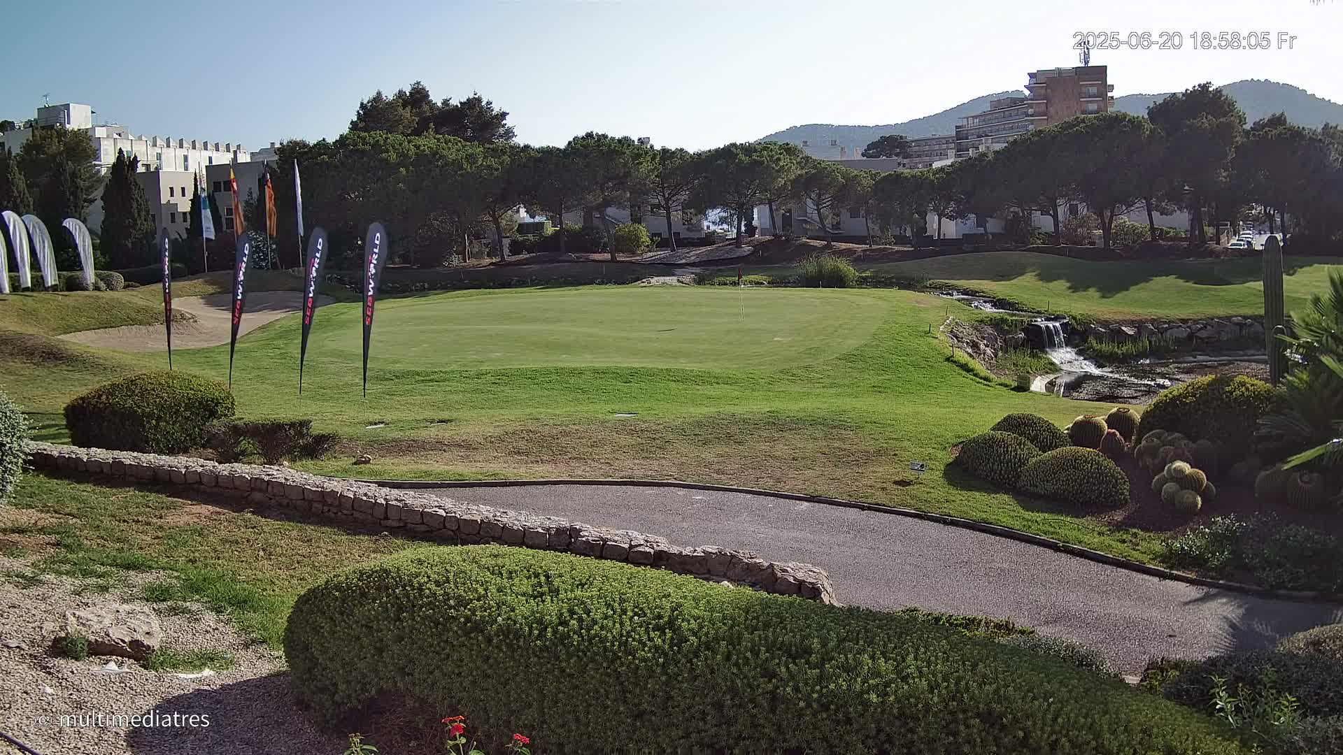 A golf course green with surrounding landscaping, a small waterfall, and buildings in the background under a clear, sunny sky.