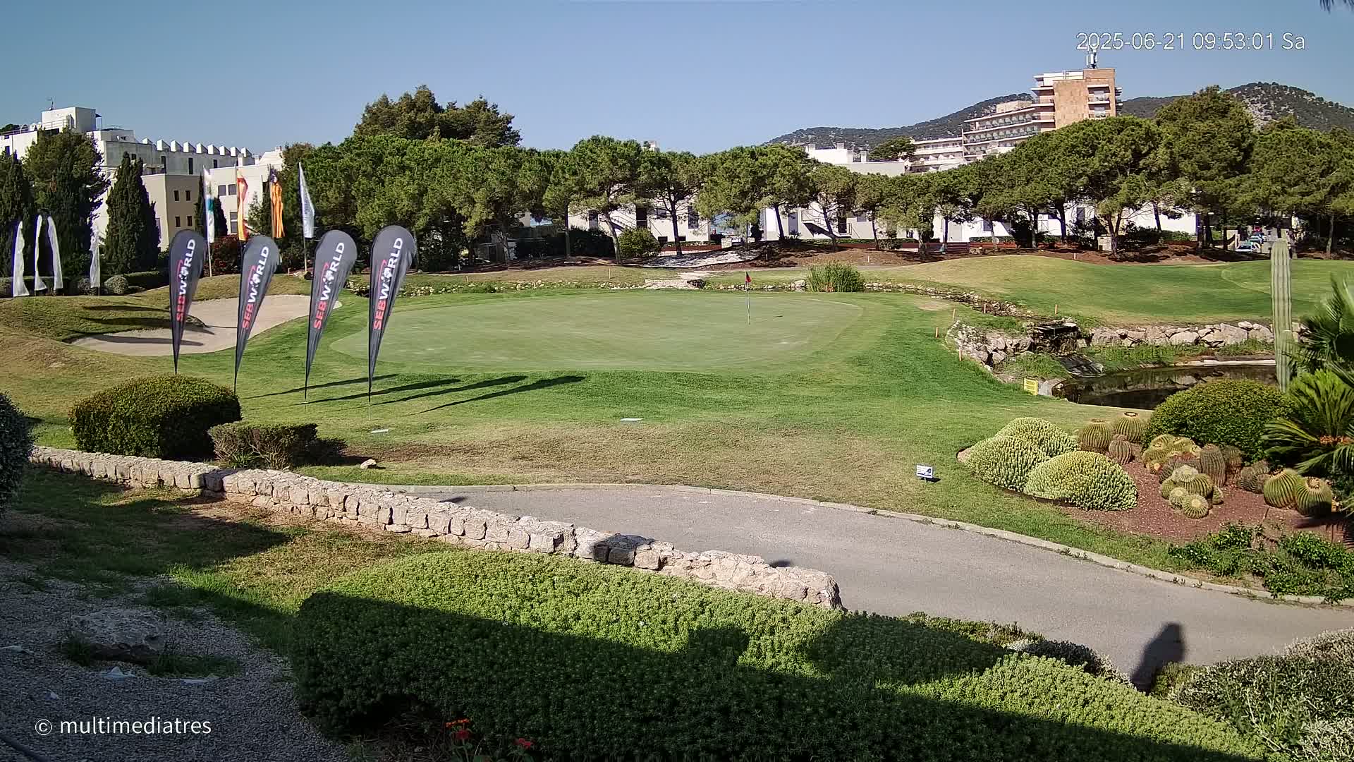 A golf course putting green with several flags, a small pond, and cacti in the foreground, under sunny skies.
