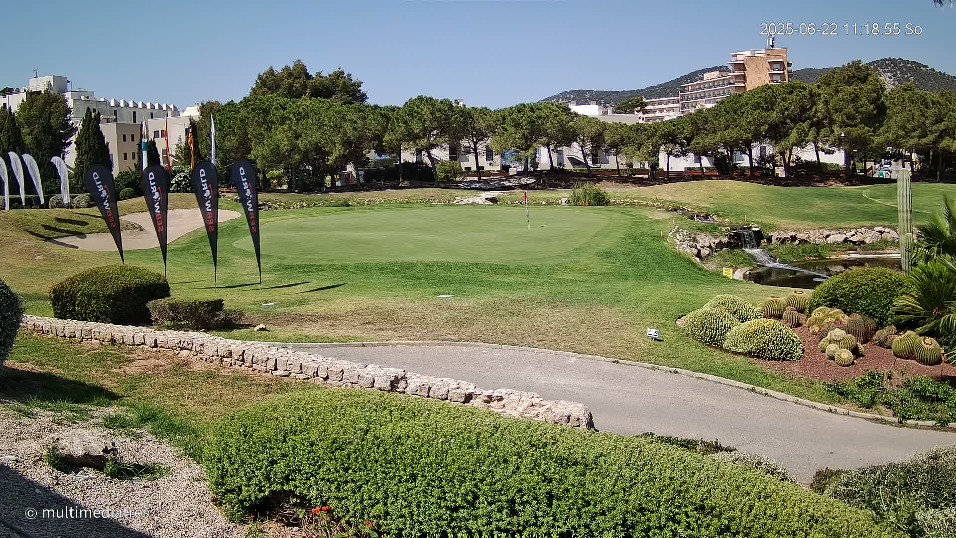 A green golf course putting green with a small waterfall and pond, surrounded by lush greenery and buildings under a clear blue sky.