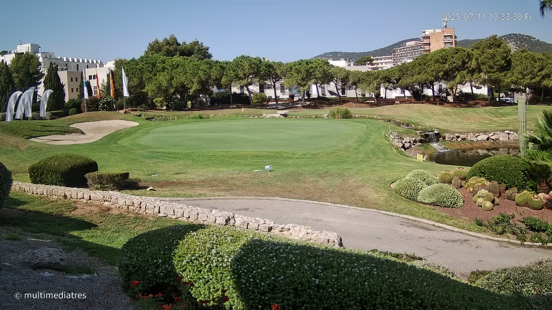 A green golf course putting green is situated near a small pond and several buildings under a clear, sunny sky.