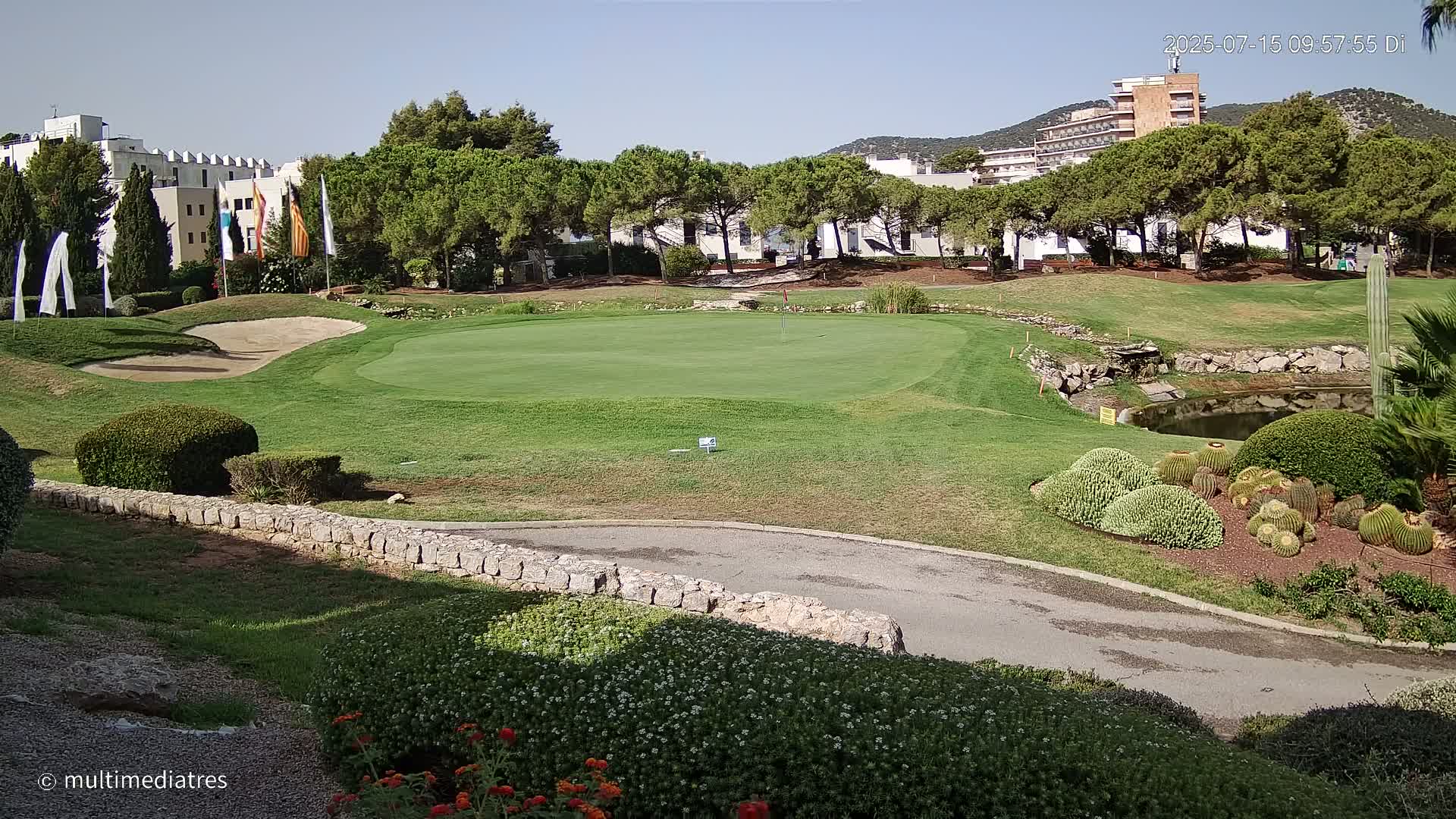 A putting green on a golf course with a sand trap and a small pond, surrounded by green trees and landscaping under a clear blue sky.