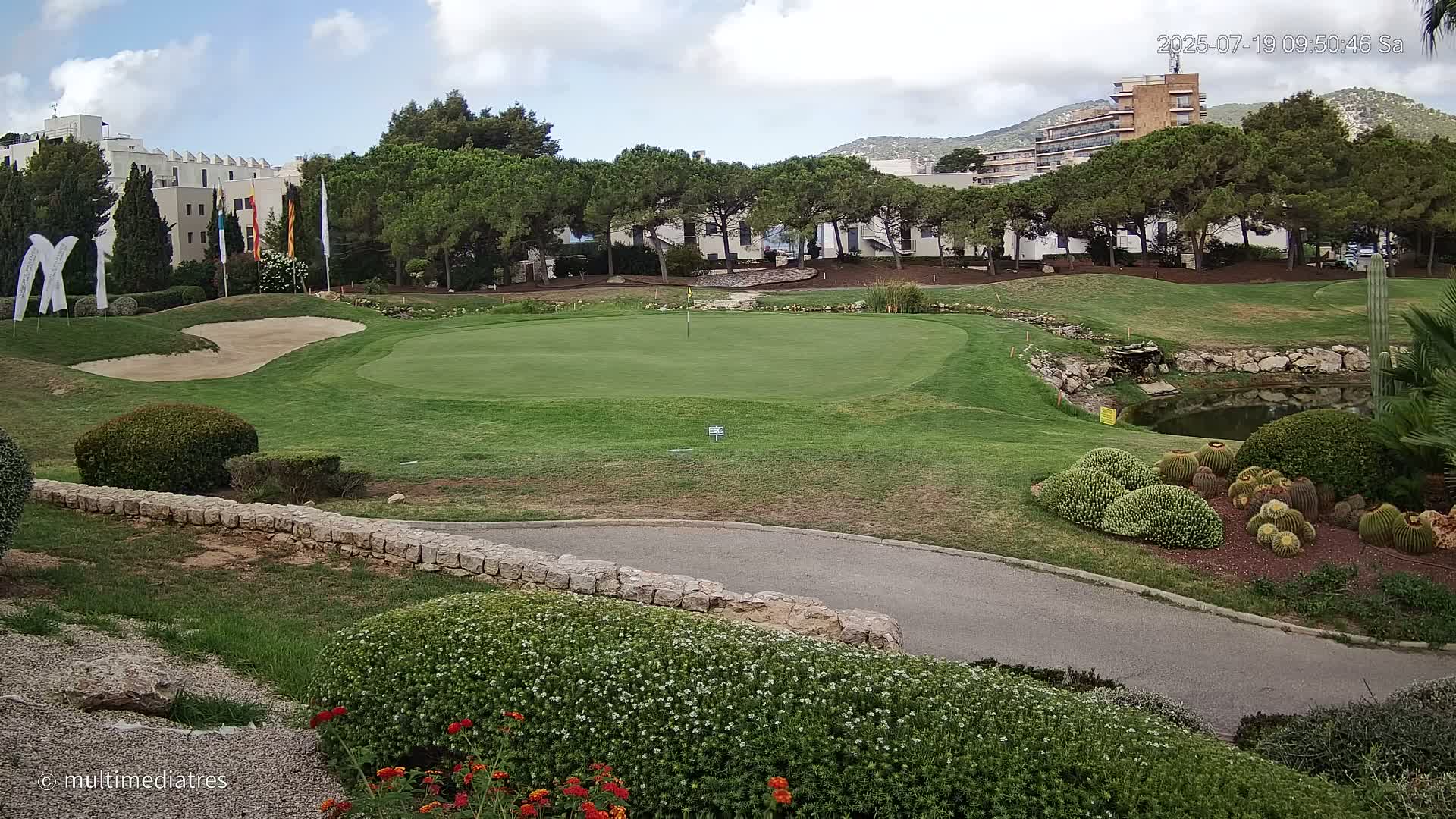 A golf course green with a sand trap and a small pond is visible under partly cloudy skies, bordered by landscaping, low stone walls, and buildings in the background.