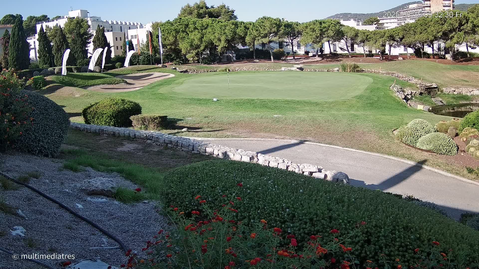 A putting green on a golf course is surrounded by landscaping, including shrubs, flowers, and a low stone wall, under a sunny sky.