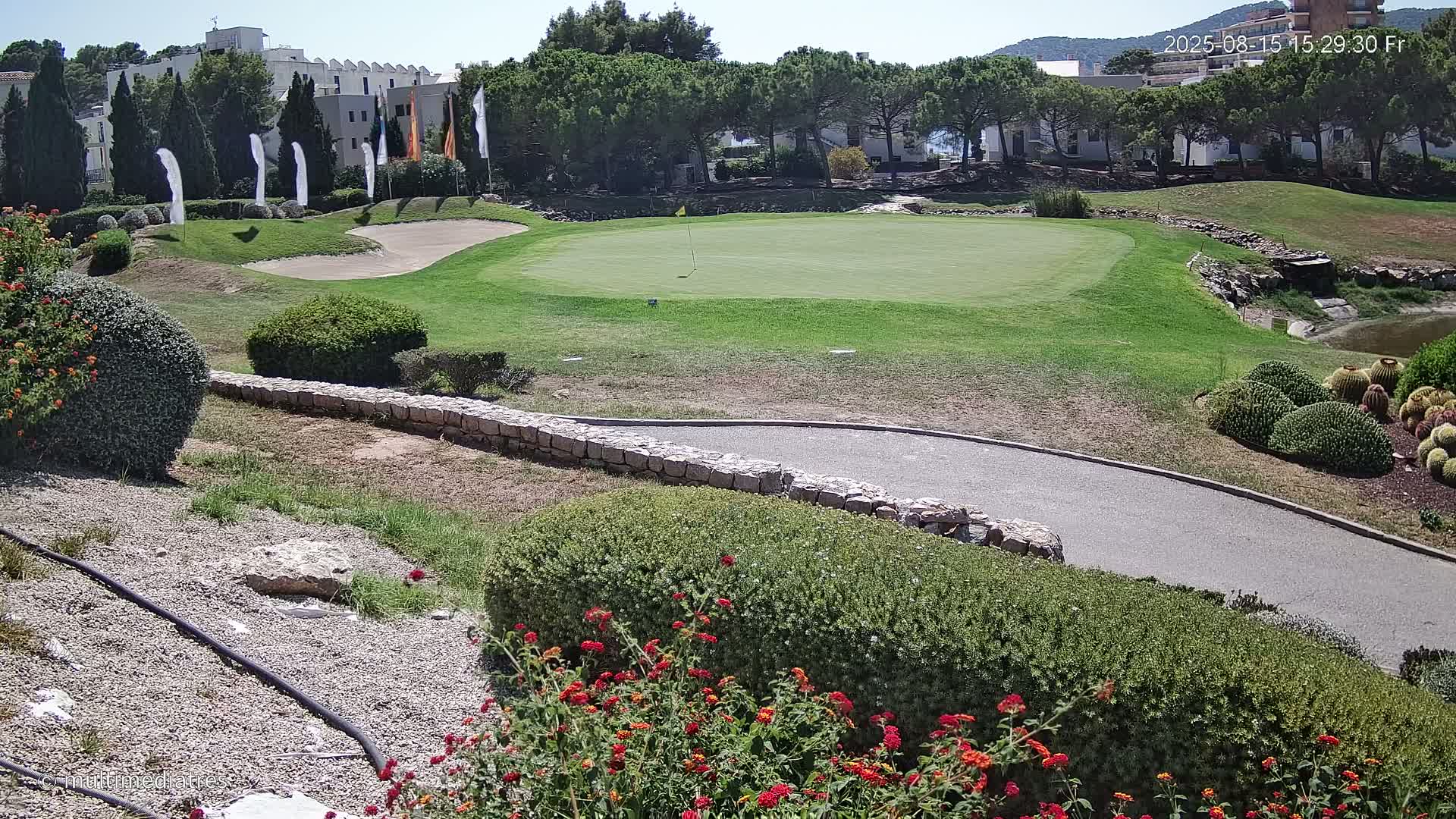 A putting green on a golf course is surrounded by landscaping, including shrubs, flowers, and a low stone wall, under a sunny sky.