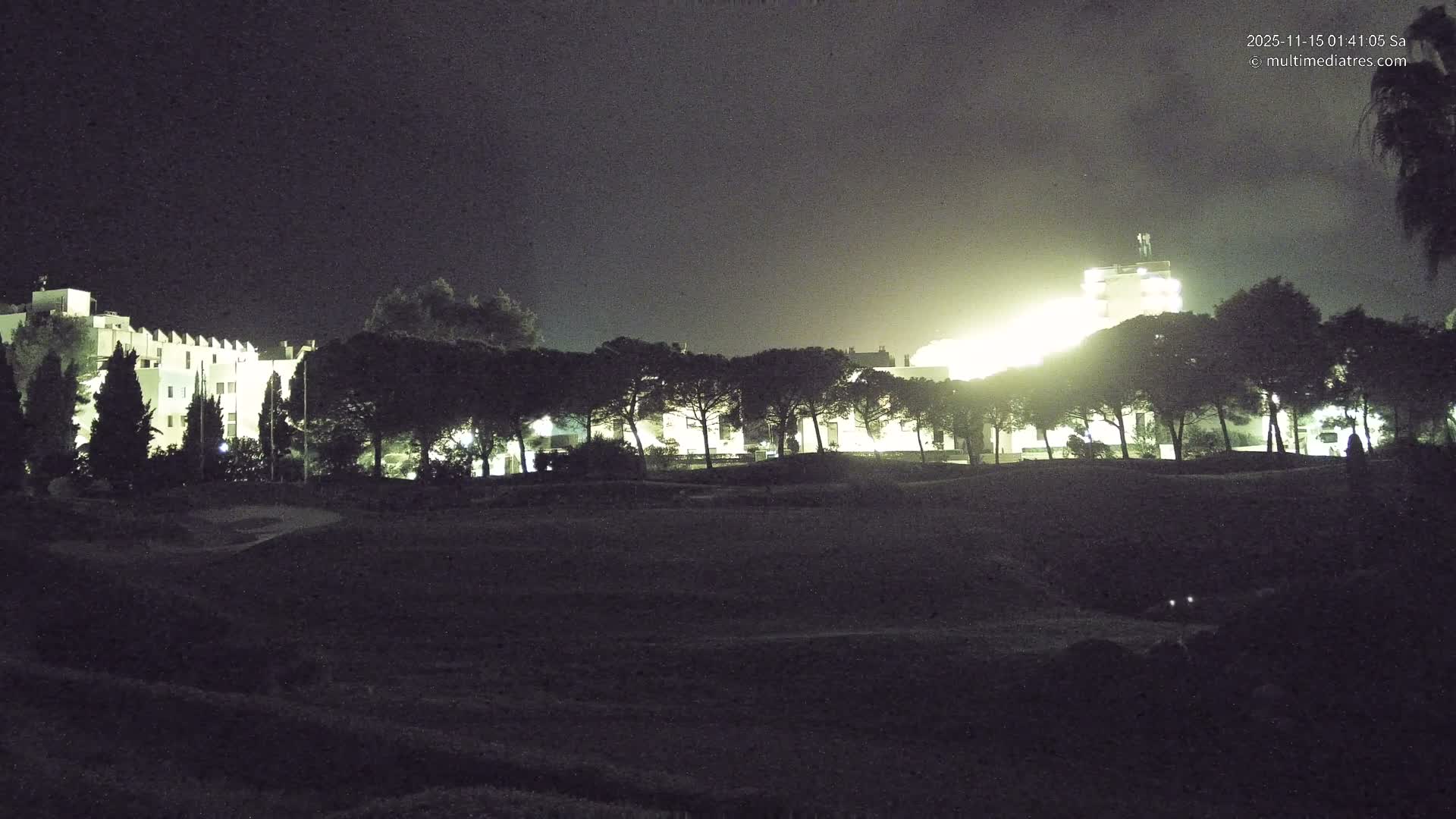 A nighttime scene reveals a dark, grassy landscape, likely a golf course, with a line of trees silhouetted against brightly lit buildings in the background, under a clear sky.