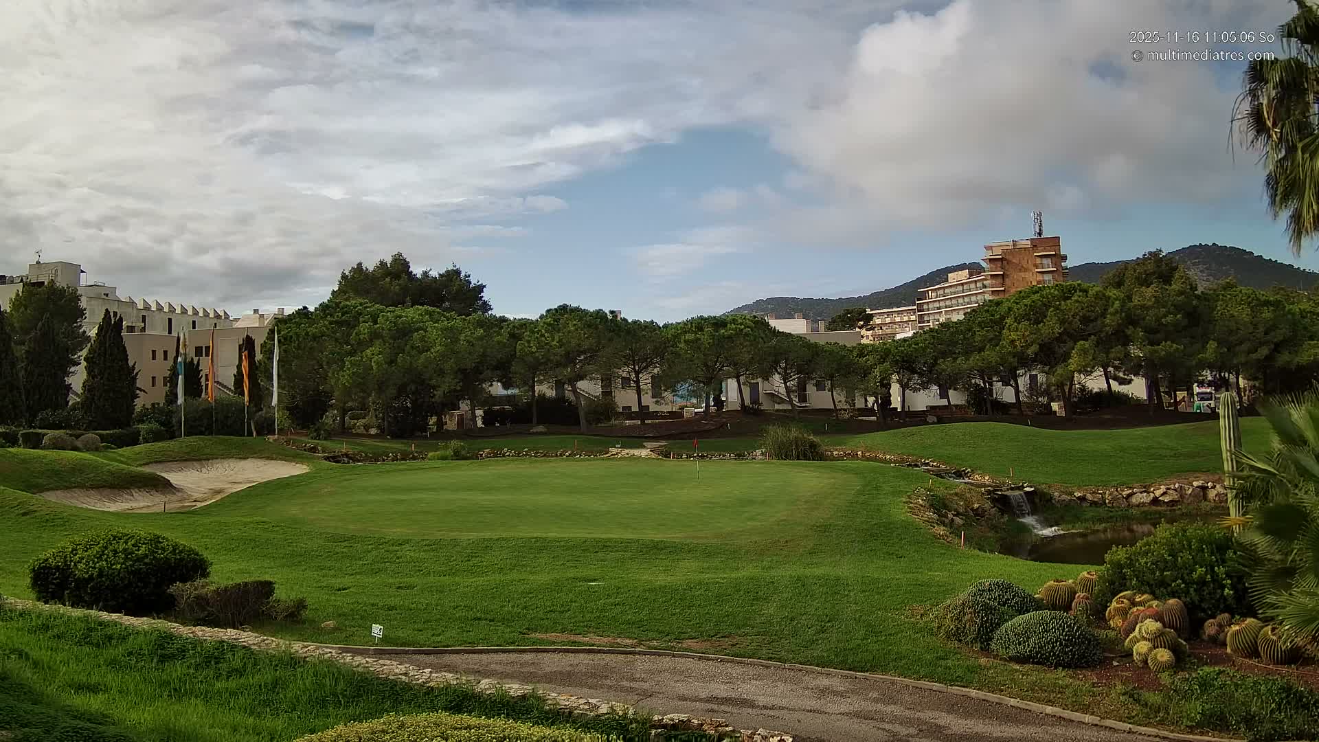 A vibrant golf course featuring a sand trap, green, and a small waterfall emptying into a pond, is framed by rows of trees, distant resort buildings, and mountains beneath a partly cloudy sky.