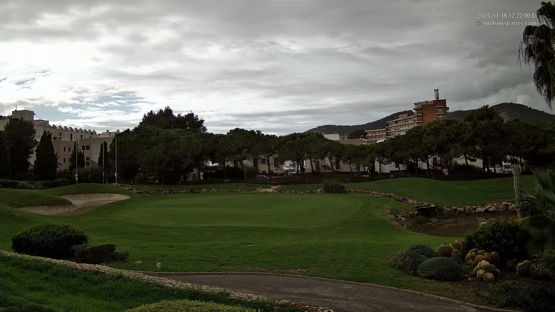 A vibrant golf course featuring a sand trap, green, and a small waterfall emptying into a pond, is framed by rows of trees, distant resort buildings, and mountains beneath a partly cloudy sky.