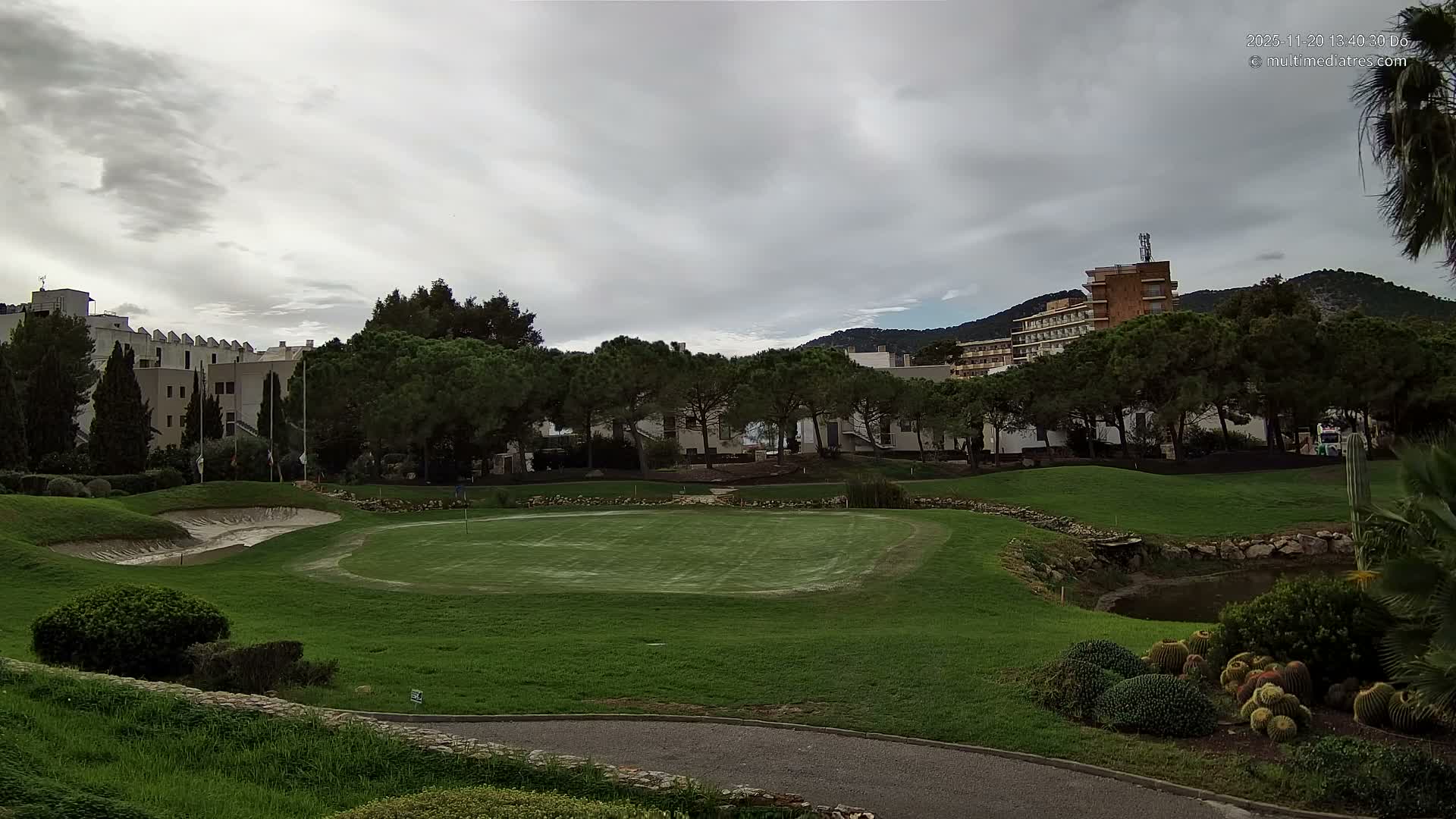 A golf course with a putting green, sand bunker, and pond is surrounded by various trees and buildings, with distant hills visible under an overcast sky.