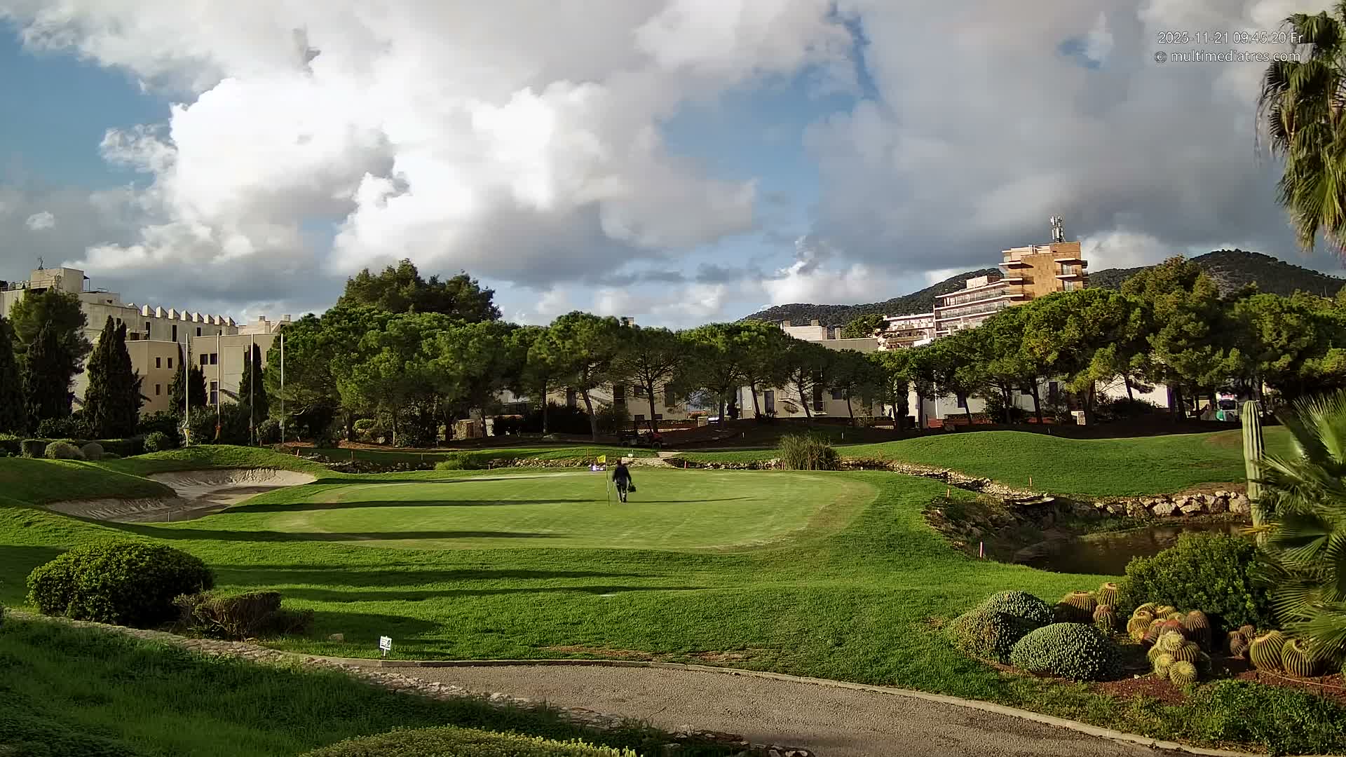 A person walks across a lush green golf course featuring a sand bunker, a small stream, and surrounded by trees, buildings, and distant hills, all under a partly cloudy sky with patches of sun creating clear shadows.