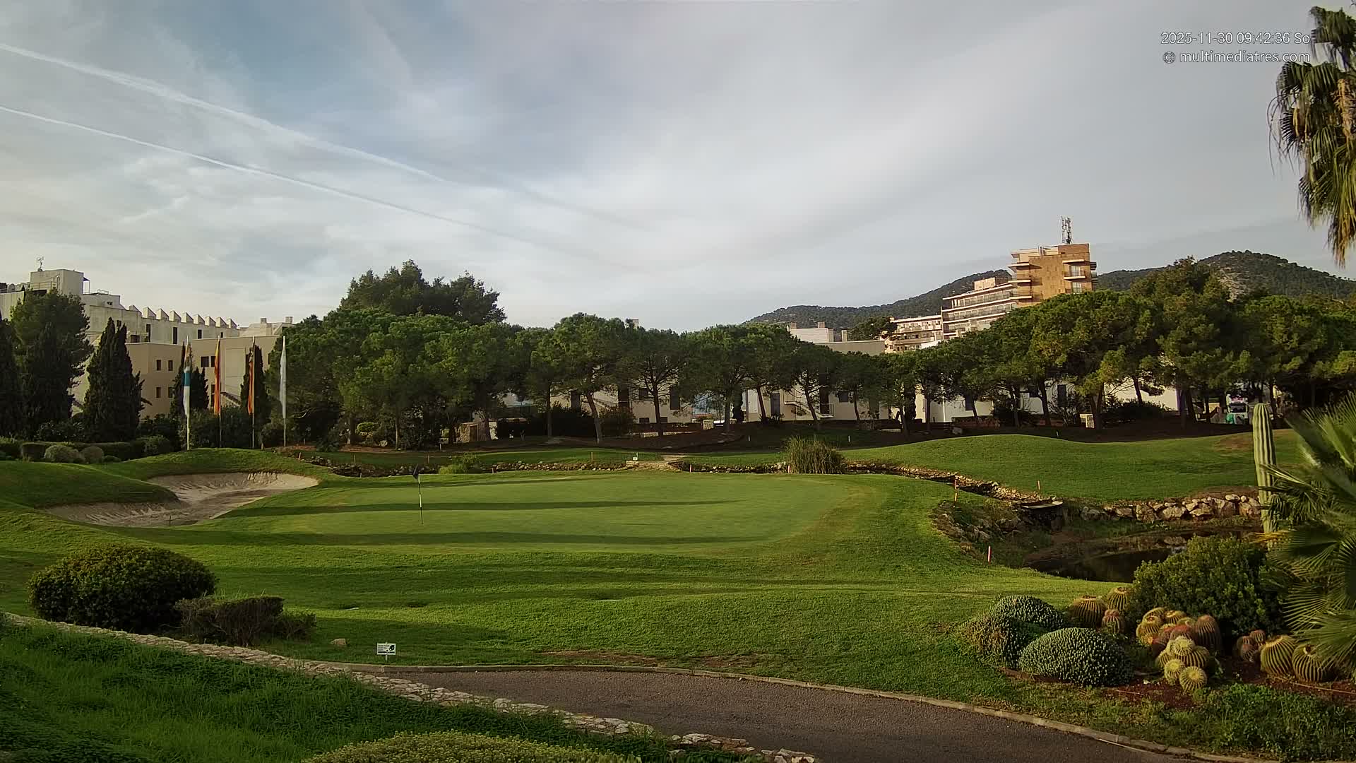 A verdant golf course featuring a sand trap, small pond, and diverse greenery including palm trees and cacti, stretches towards various buildings and distant hills under a partly cloudy sky.
