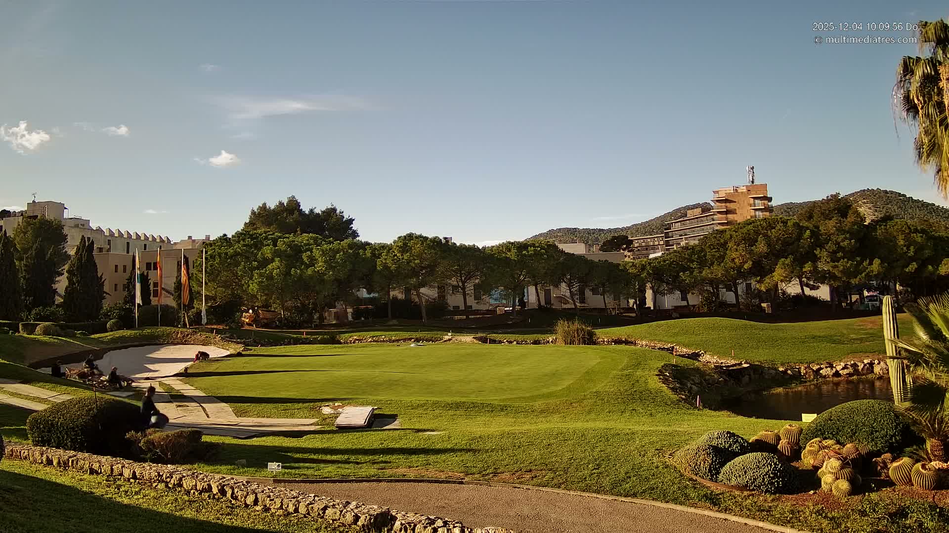 A sunny day with a clear blue sky and wispy clouds illuminates a verdant golf course with bunkers, a pond, and numerous trees, set against resort buildings and distant mountains, with a few people working on the course.