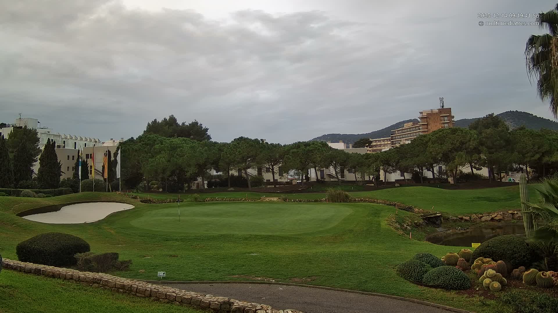 A sunny day with a clear blue sky and wispy clouds illuminates a verdant golf course with bunkers, a pond, and numerous trees, set against resort buildings and distant mountains, with a few people working on the course.