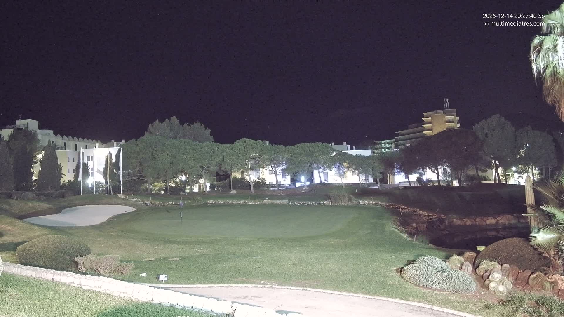 A sunny day with a clear blue sky and wispy clouds illuminates a verdant golf course with bunkers, a pond, and numerous trees, set against resort buildings and distant mountains, with a few people working on the course.