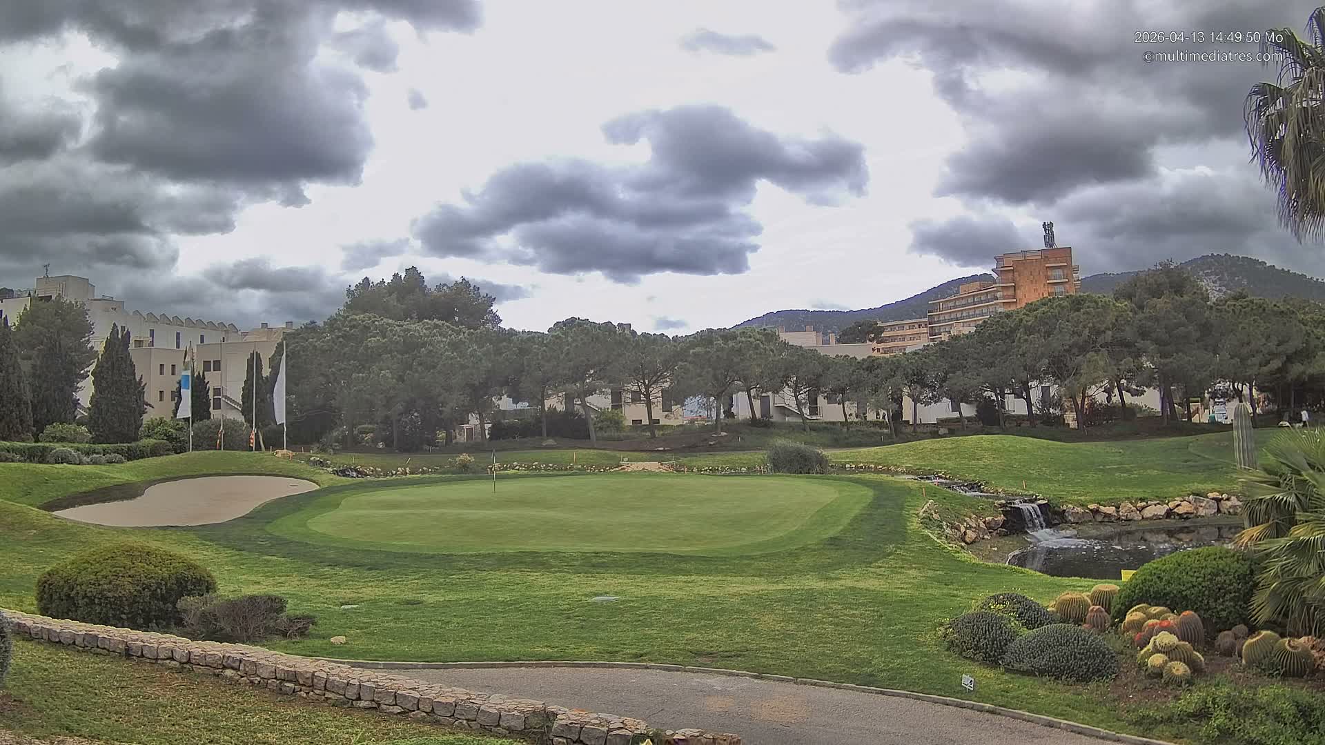 A sunny day with a clear blue sky and wispy clouds illuminates a verdant golf course with bunkers, a pond, and numerous trees, set against resort buildings and distant mountains, with a few people working on the course.