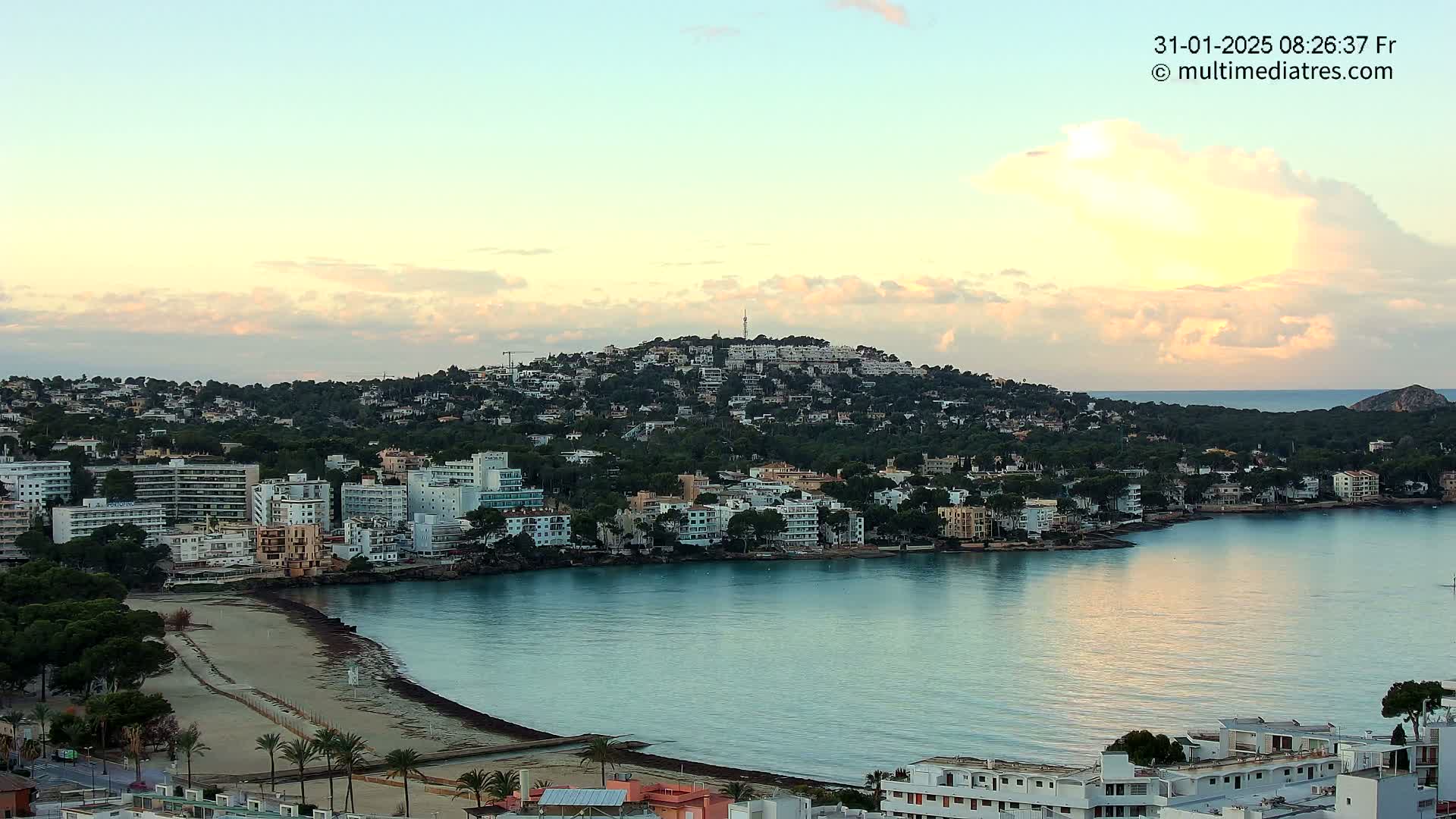 A coastal town nestled against a hill, with calm waters reflecting the pale sunrise sky.