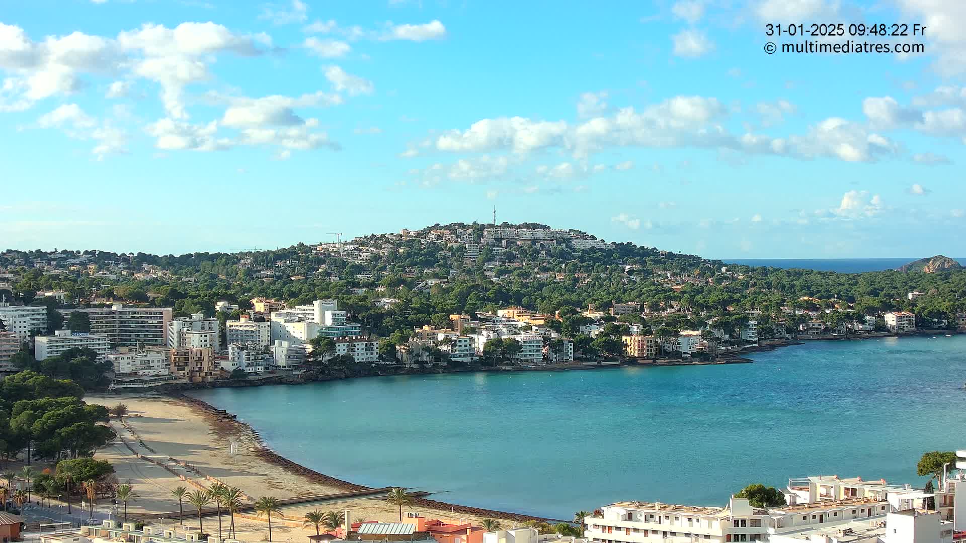 A coastal town nestled against a verdant hill overlooking a calm, turquoise bay under a mostly sunny sky with scattered clouds.