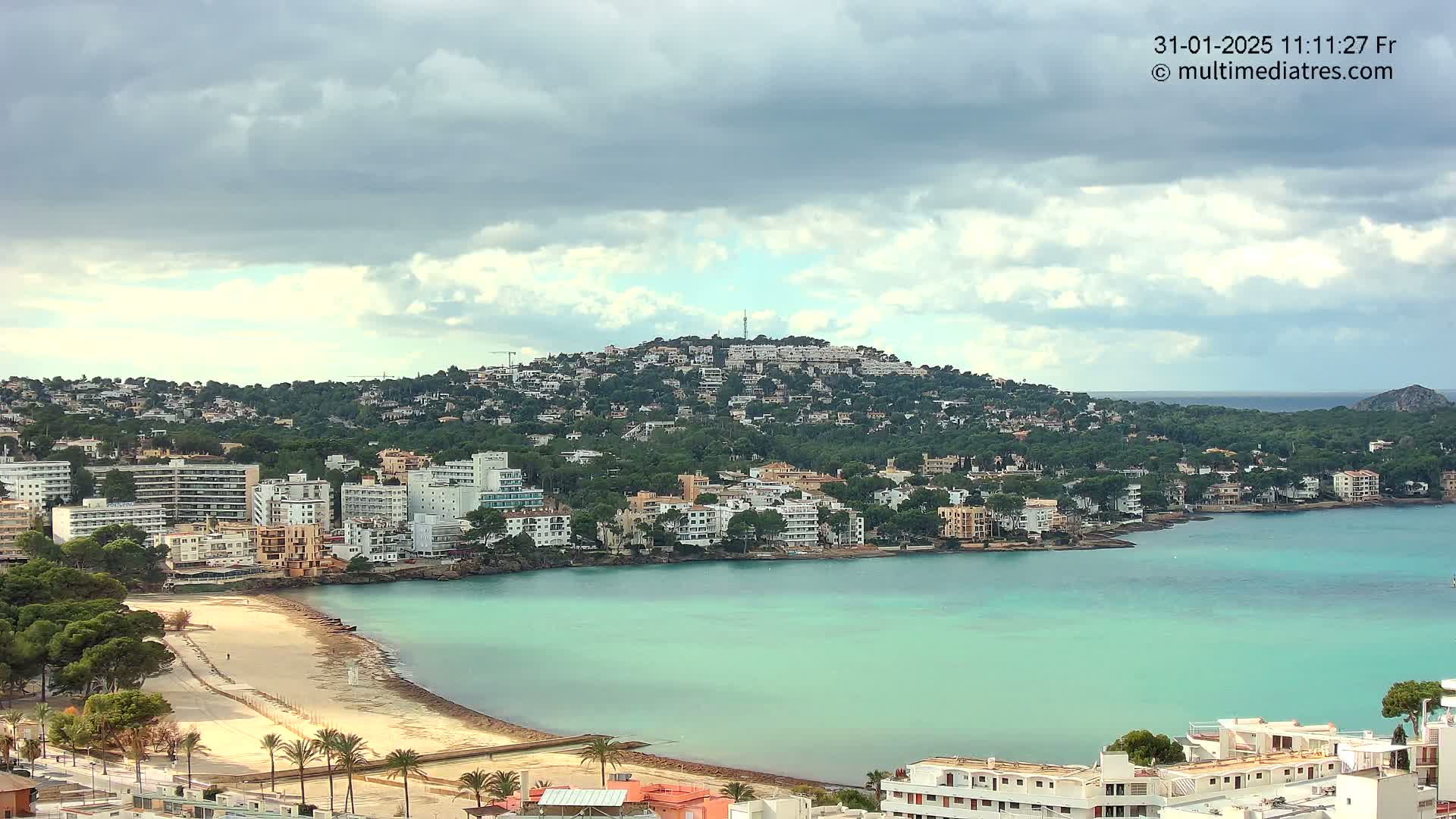 A coastal town with light-colored buildings clusters around a calm, teal bay under a partly cloudy sky.