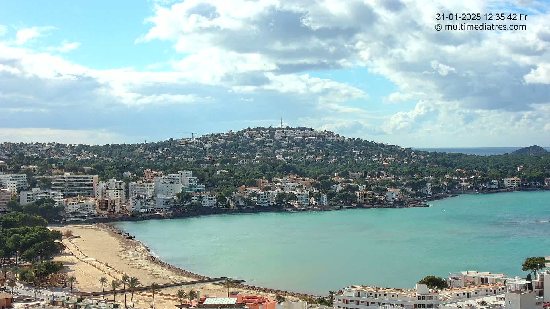 A coastal town with a sandy beach and turquoise water sits nestled below a hill covered in houses and greenery under a partly cloudy sky.