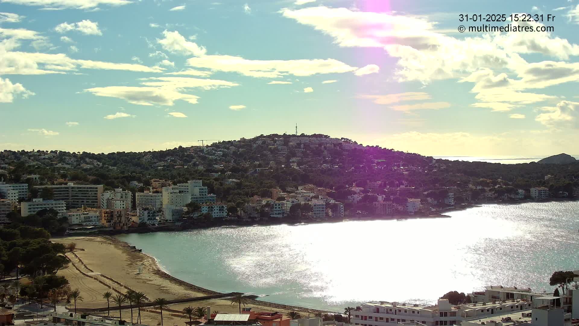 A sunny day reveals a coastal town nestled against a hill, with a calm bay reflecting sunlight.