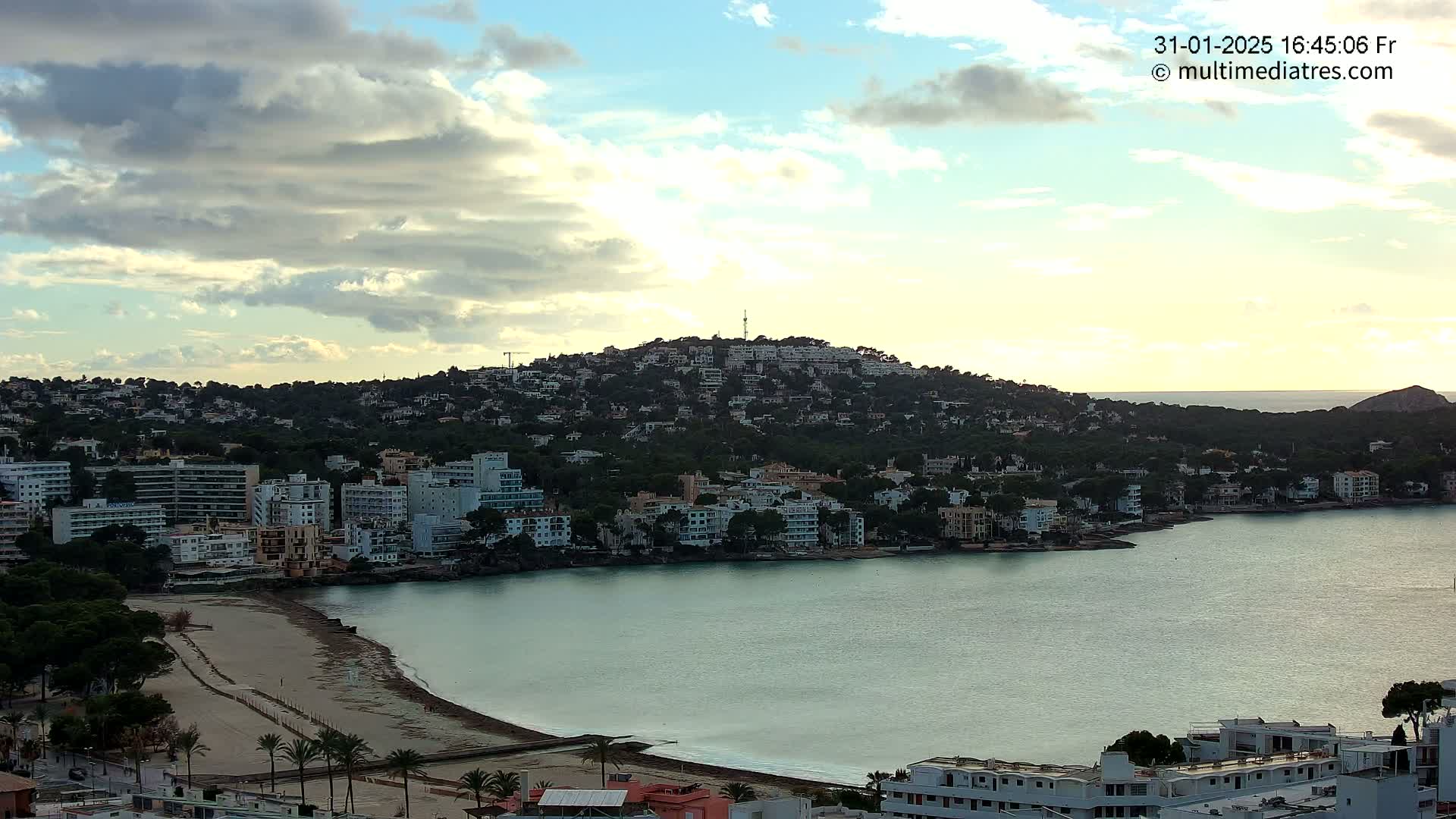 A coastal town nestled against a hill, under a partly cloudy sky, features a calm bay with a sandy beach.