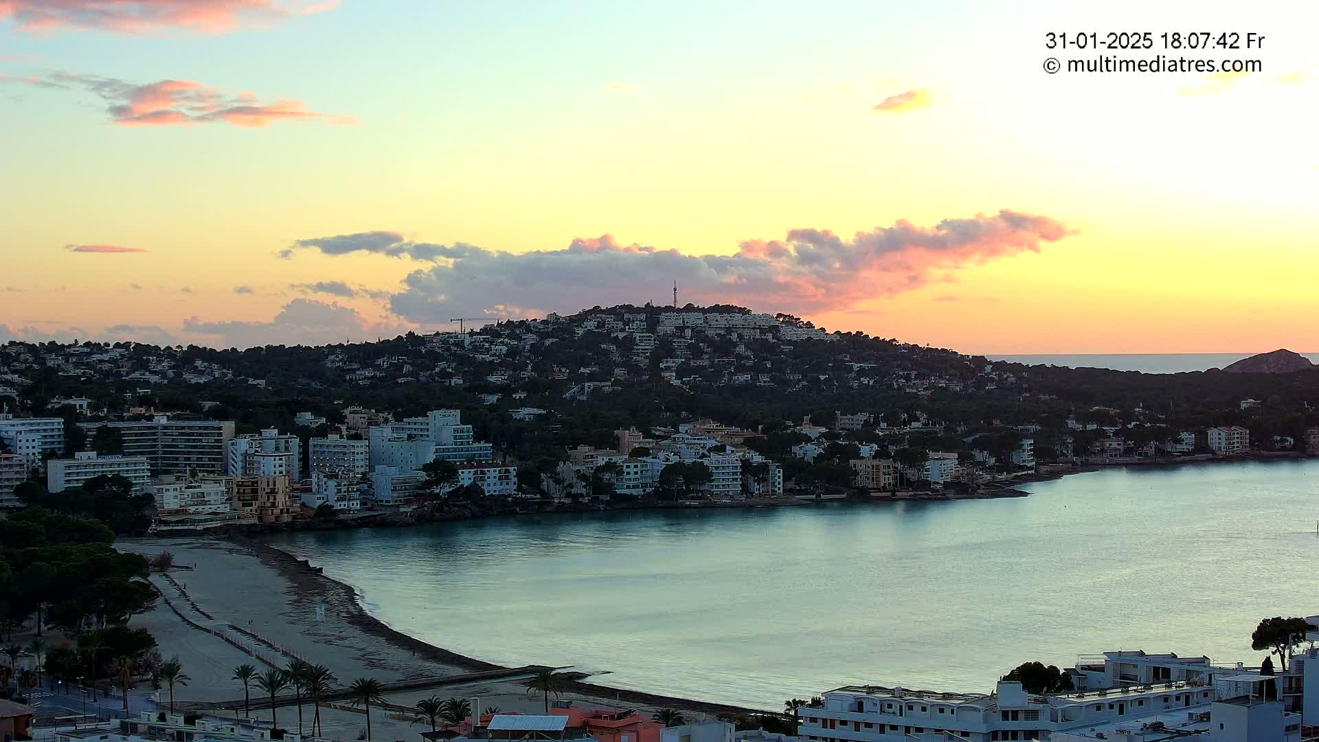 A coastal town with buildings covering a hill, a sandy beach, and calm water, all under a sunset sky with pink and orange clouds.