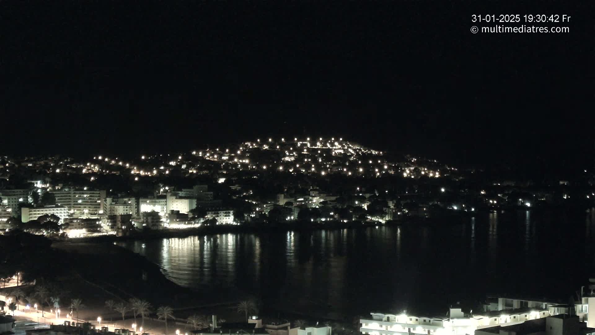A nighttime view of a coastal town with many lights illuminating buildings along a calm body of water.