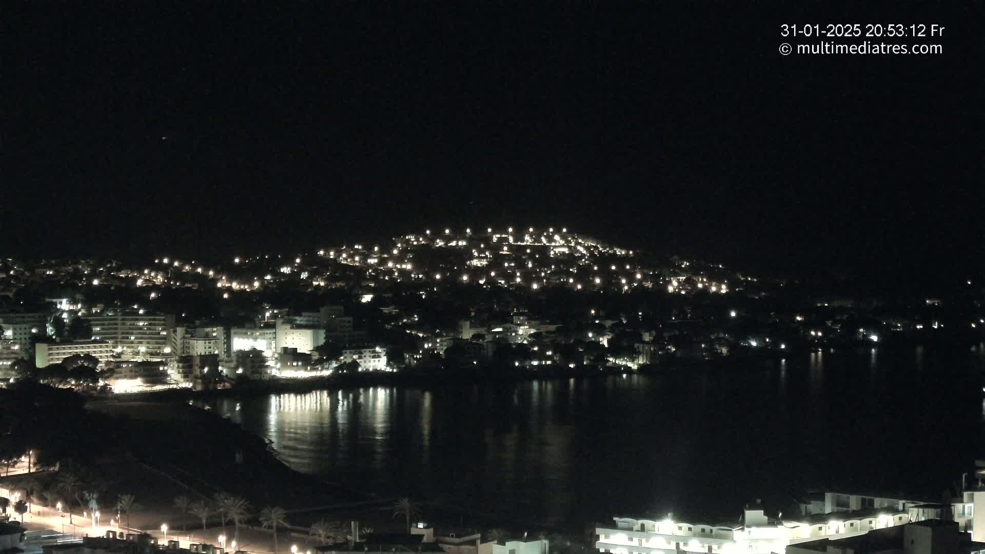 A nighttime view of a coastal town illuminated by lights, reflecting on a calm body of water.