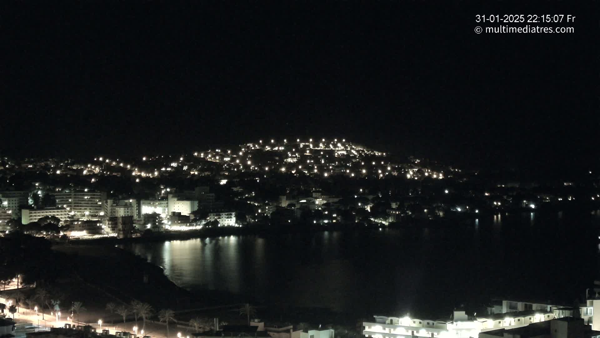 A nighttime view of a coastal town illuminated by numerous lights, with calm water reflecting the city lights.