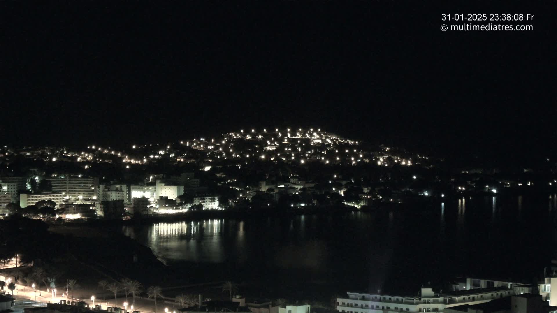 A nighttime view of a coastal town illuminated by lights, with its reflection visible in a calm body of water.