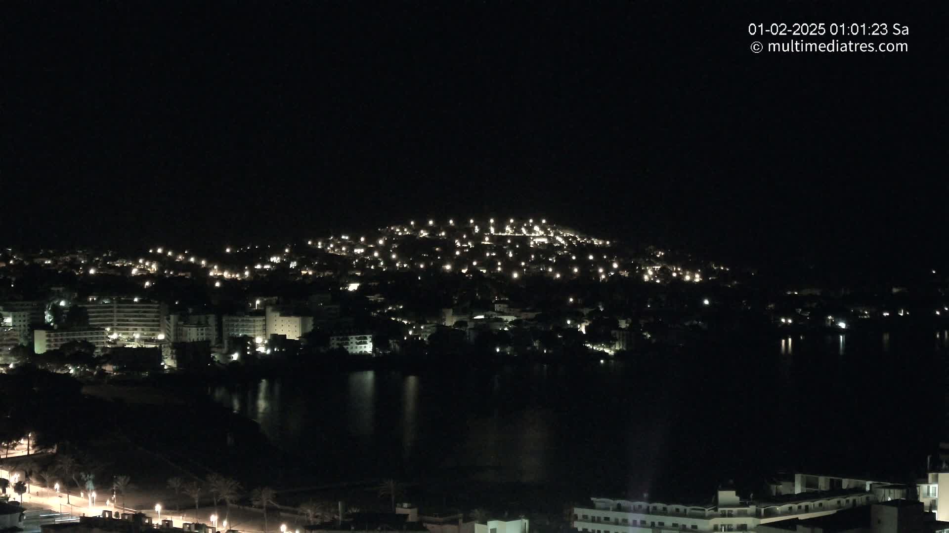 A nighttime view of a city illuminated by many lights, situated beside a dark body of water under a clear, dark sky.