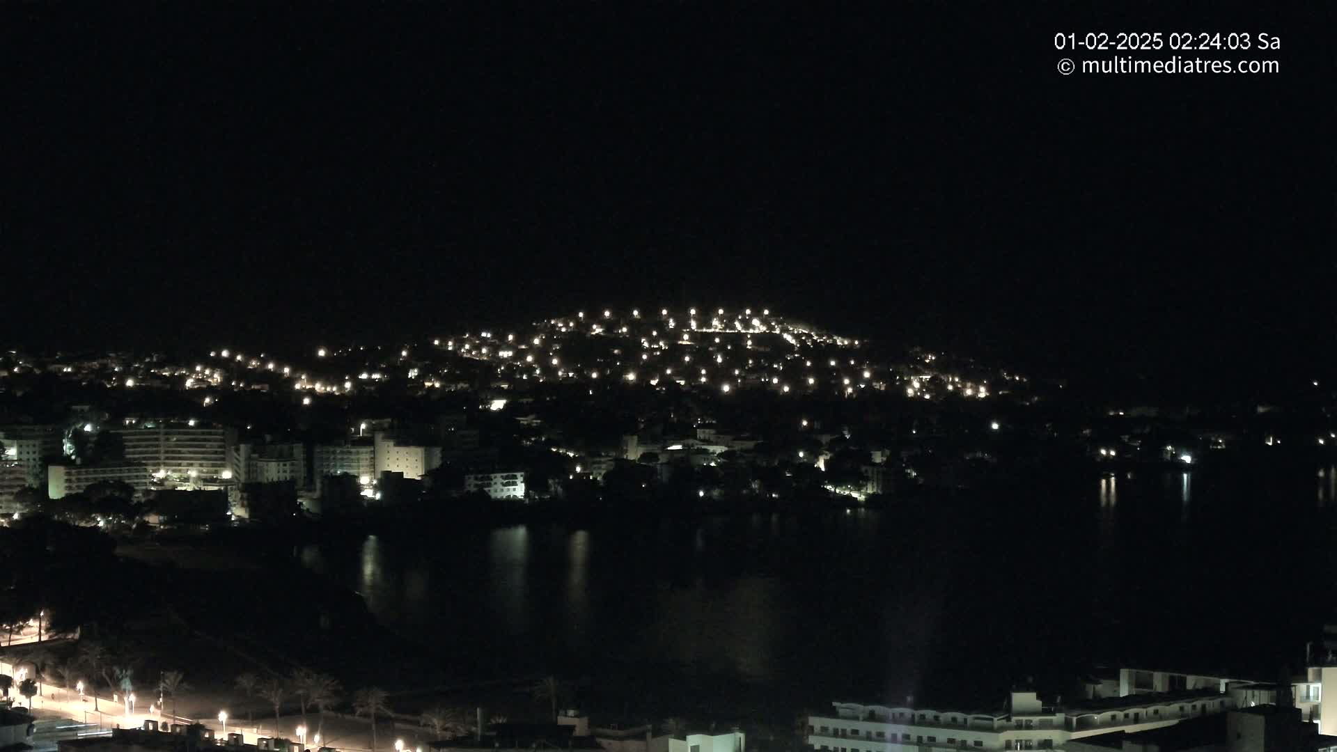 A nighttime view of a city illuminated by lights, situated beside a dark body of water, under a clear, dark sky.
