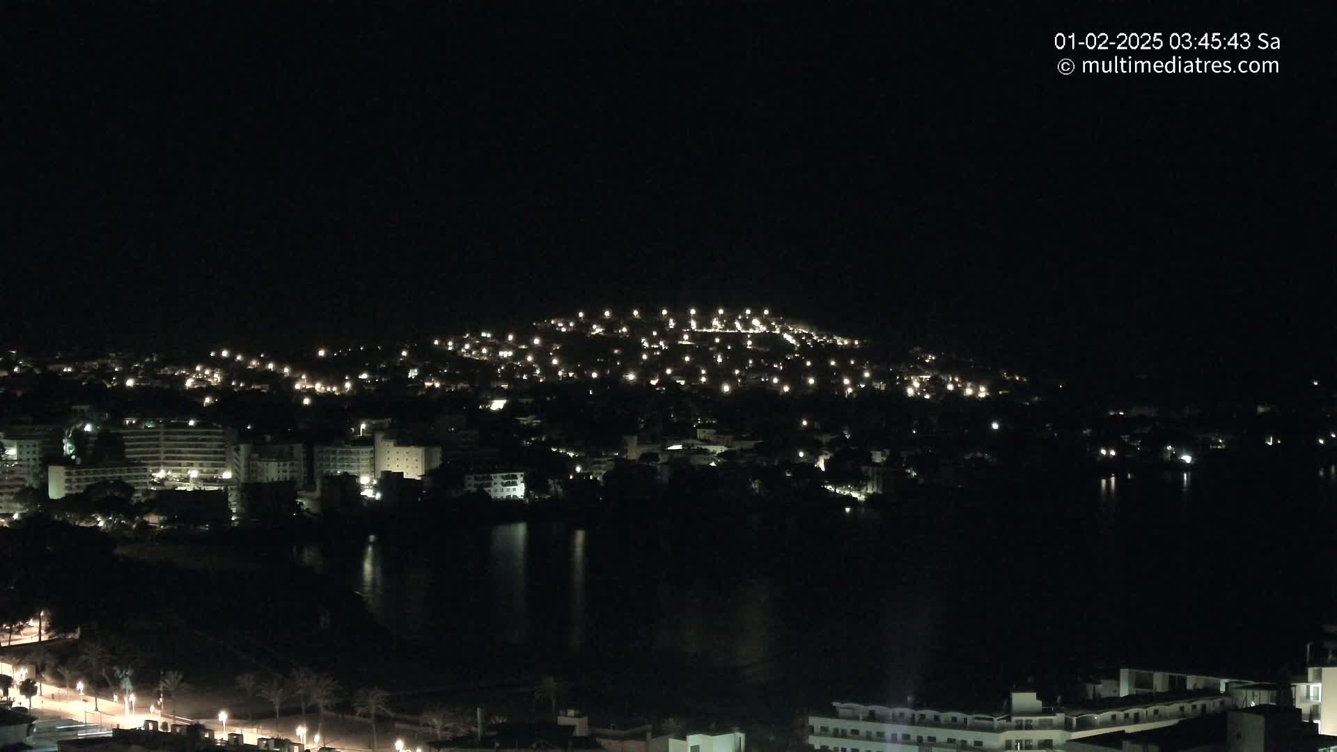 A nighttime view of a city illuminated by lights, situated alongside a dark body of water, with a hill in the background also speckled with lights.