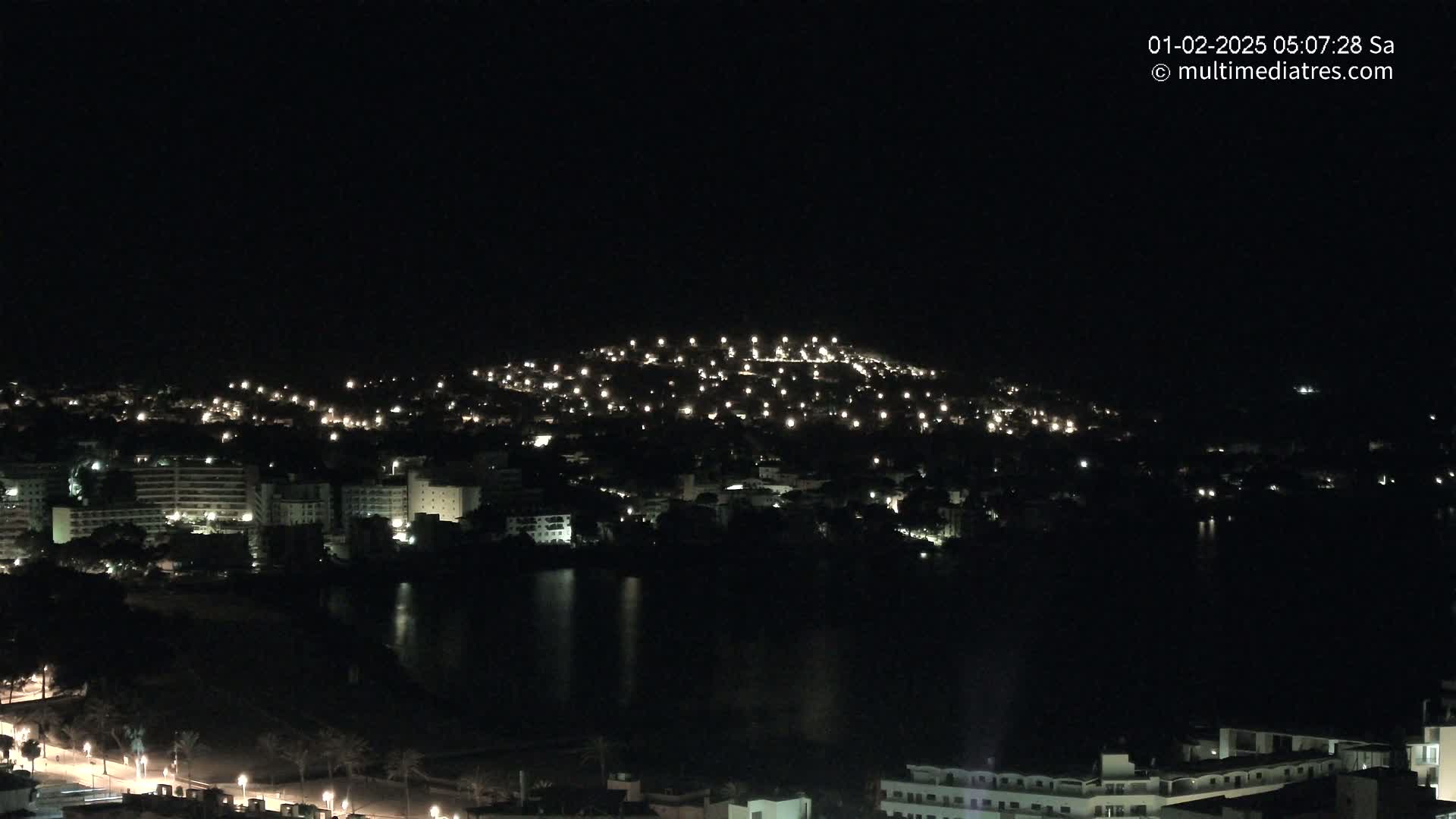 A nighttime view of a city spread across a hill and along a dark body of water, illuminated by scattered lights.