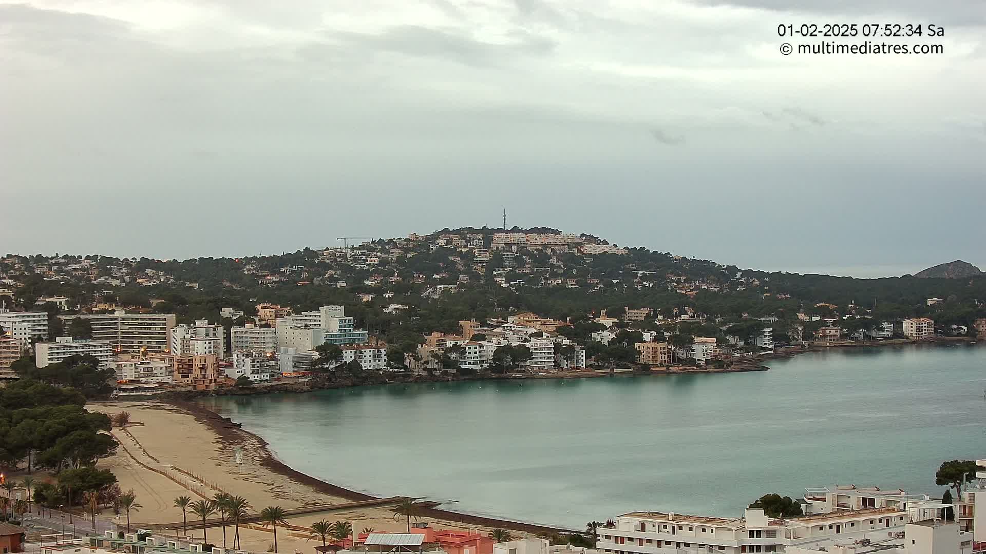 A coastal town nestled against a hill under an overcast sky overlooks a calm bay with a sandy beach.