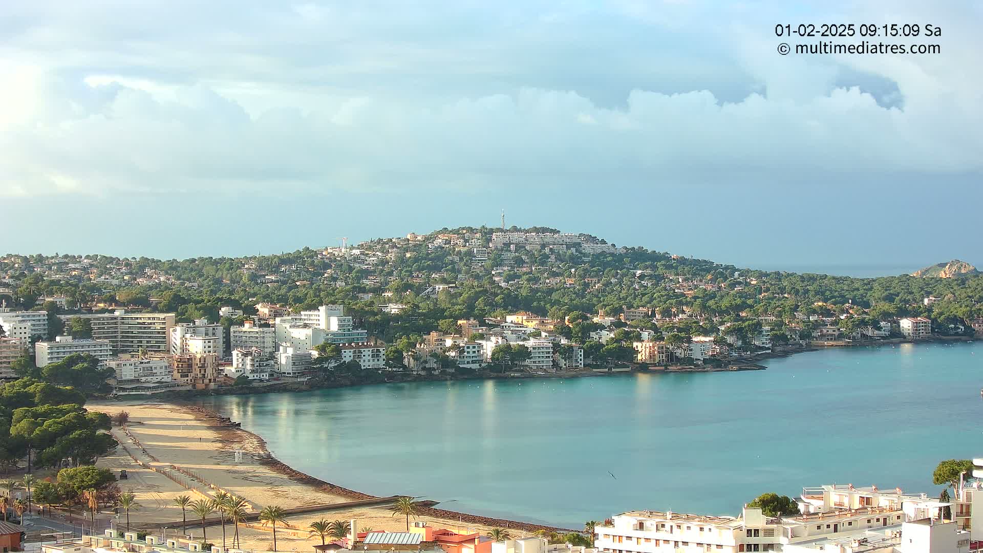 A coastal town nestled against a hill, with a sandy beach and calm, blue water under a partly cloudy sky.