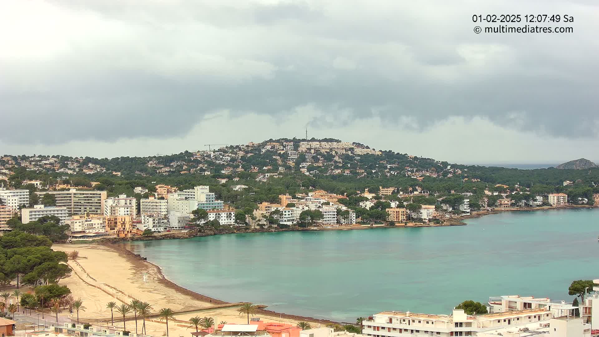 A coastal town with numerous buildings and a sandy beach is nestled below a hill covered in vegetation under an overcast sky.