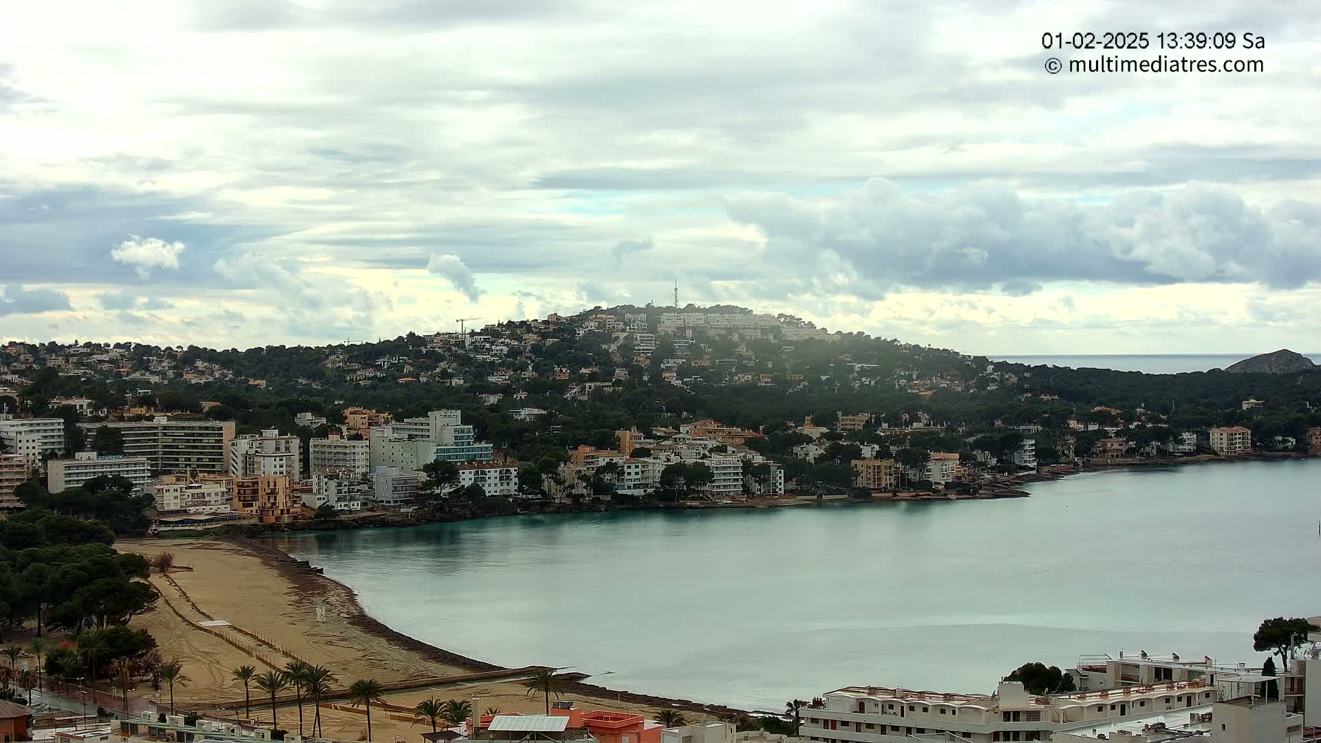 A coastal town nestled on a hillside under a mostly cloudy sky overlooks a calm bay with a sandy beach.