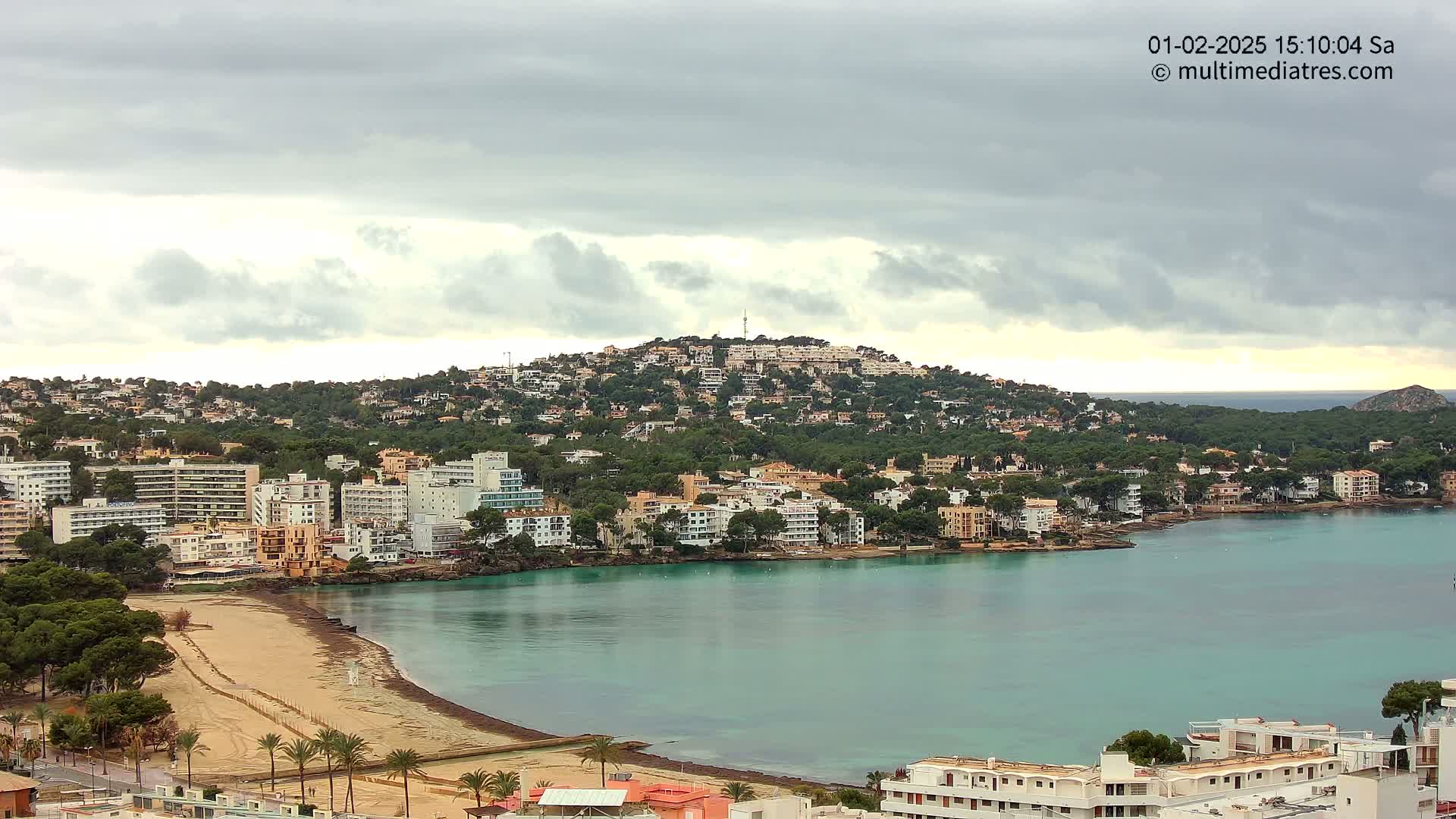 A coastal town with numerous buildings nestled against a green hill overlooks a calm, teal bay under a cloudy sky.