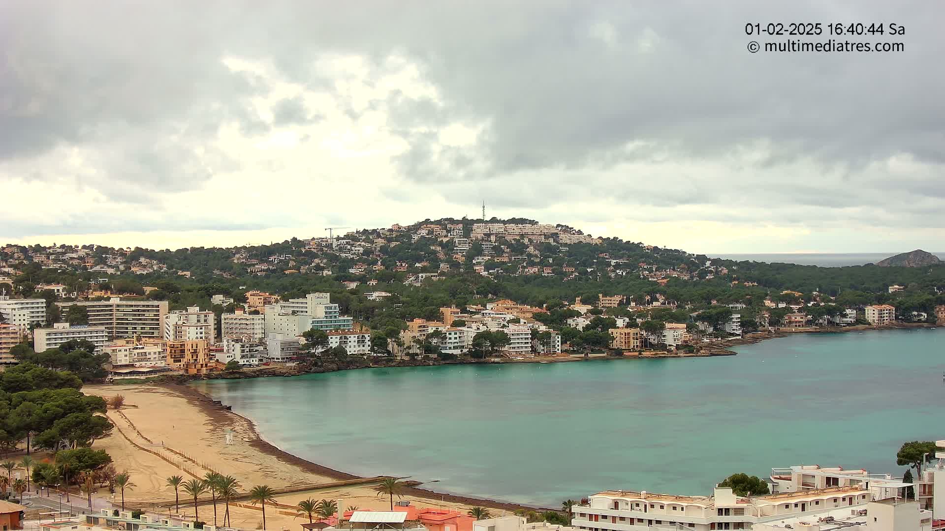 A coastal town with buildings situated on a hillside and along a calm, teal bay under a cloudy sky.
