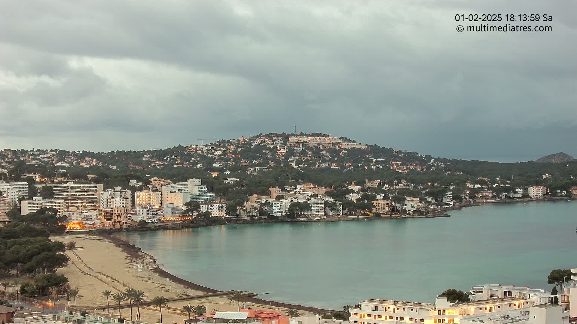 A coastal town with buildings illuminated at dusk under a cloudy sky, nestled around a calm bay with a sandy beach.