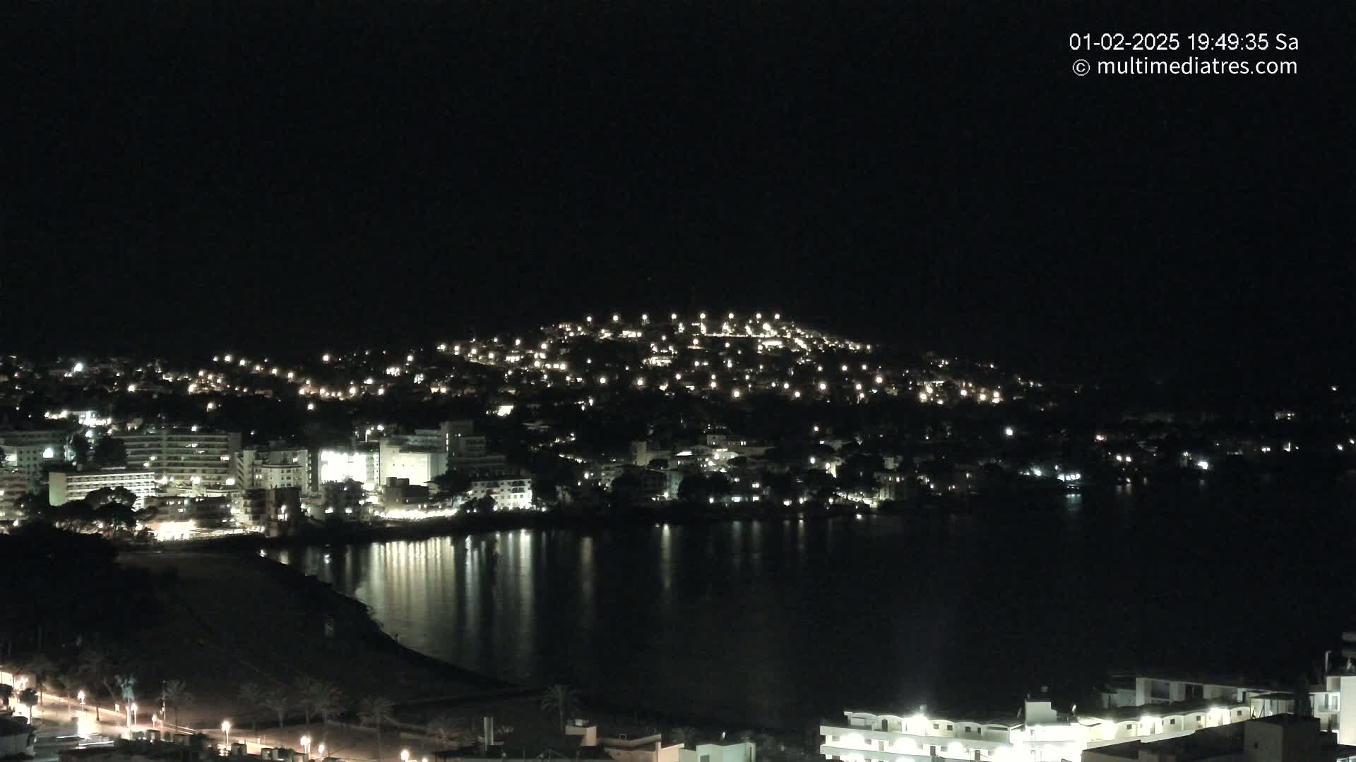 A nighttime view of a coastal town illuminated by lights, with its buildings clustered on a hill overlooking a calm, dark body of water.
