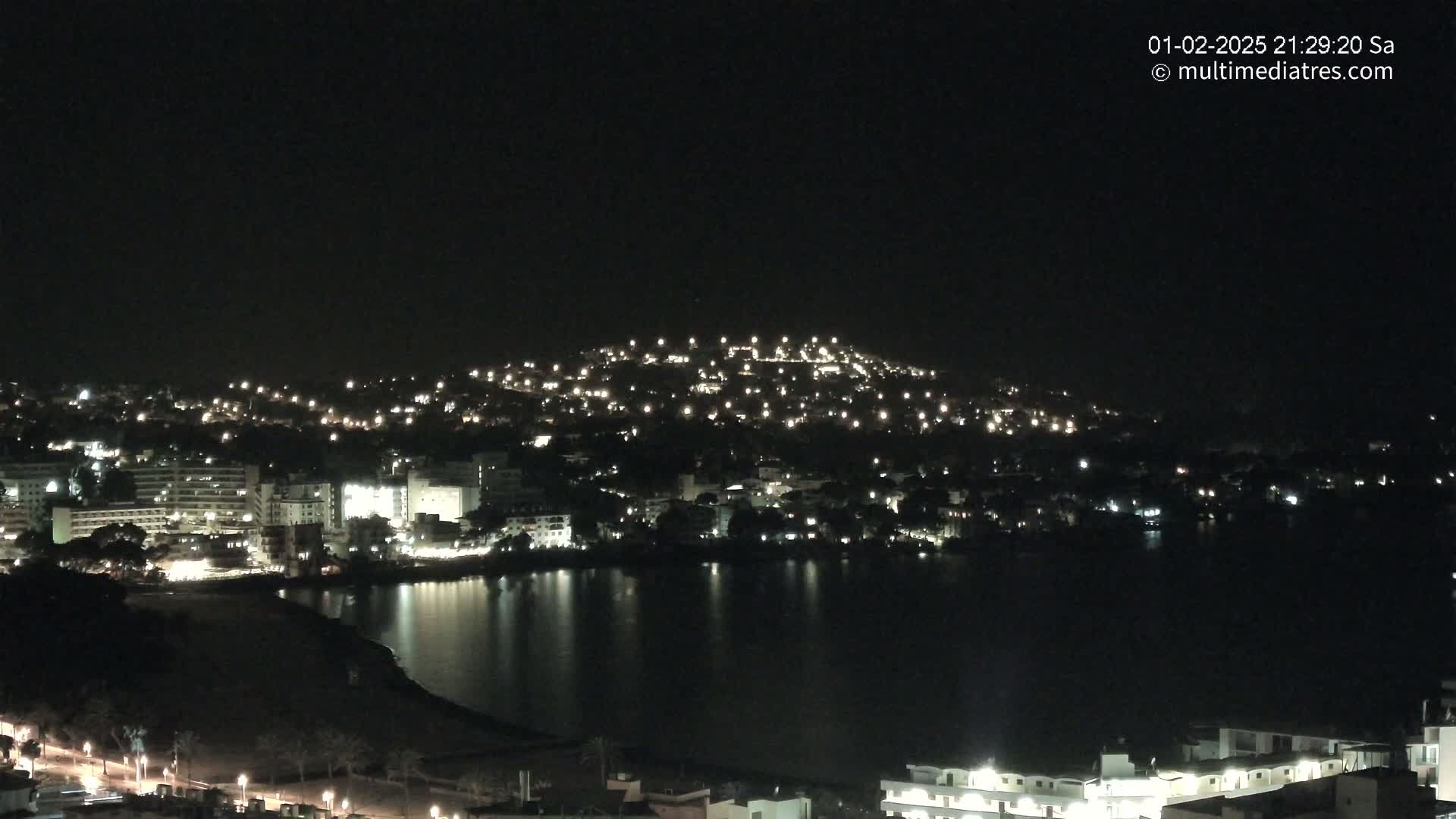 A nighttime view of a coastal town with many lights illuminating buildings and streets, reflected in a calm body of water.
