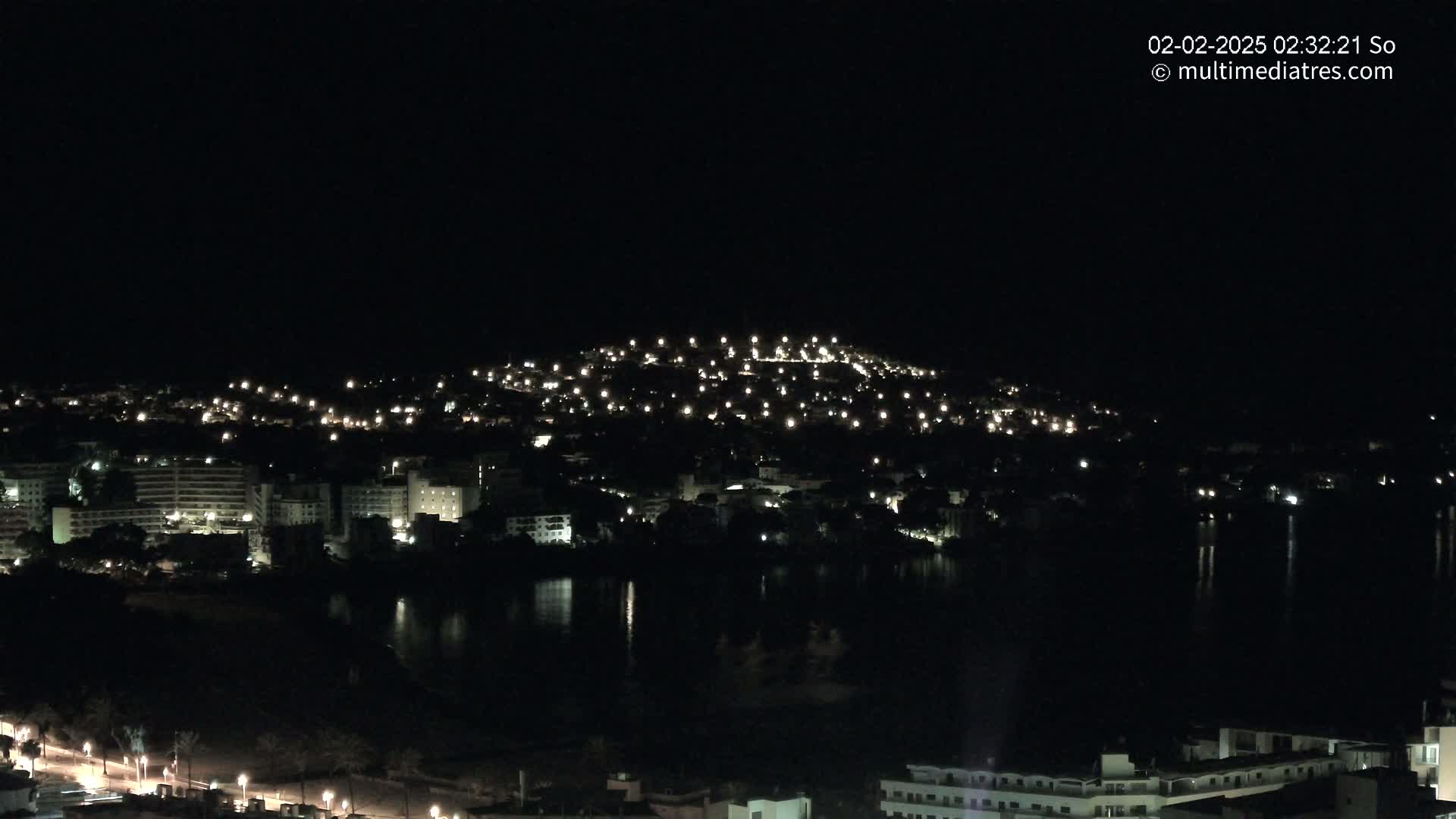 A nighttime view of a town situated on a hill next to a body of water, illuminated by numerous lights.