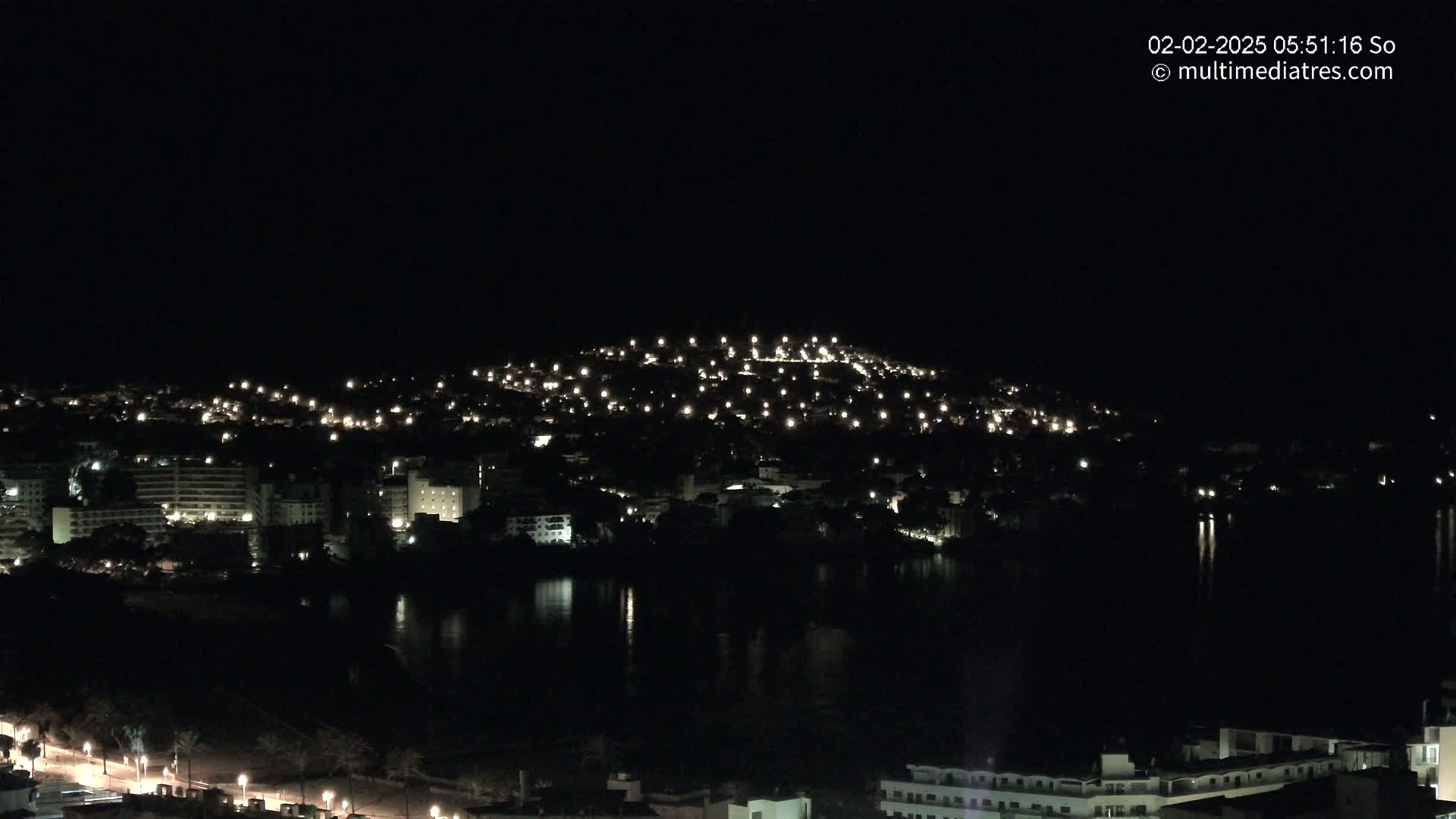 A coastal town at night, illuminated by many small lights, is reflected in a dark body of water under a clear, dark sky.